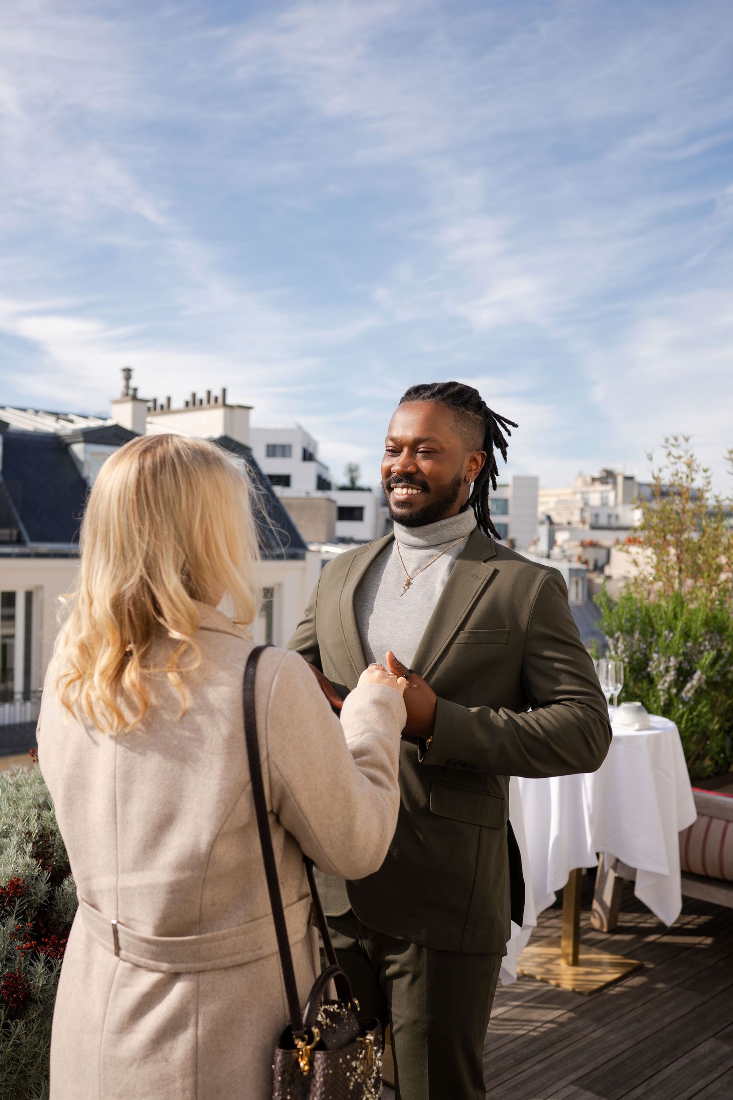 A smiling man in an olive suit holds hands with a blonde woman on a Parisian rooftop terrace under a blue sky.