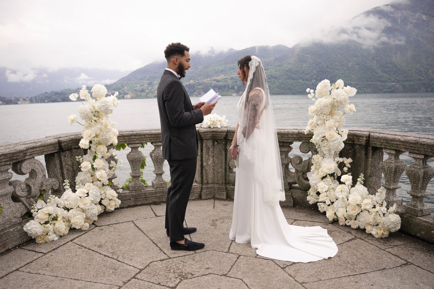 Couple exchanging vows on elegant lakeside terrace adorned with white floral arrangements and mountain backdrop.