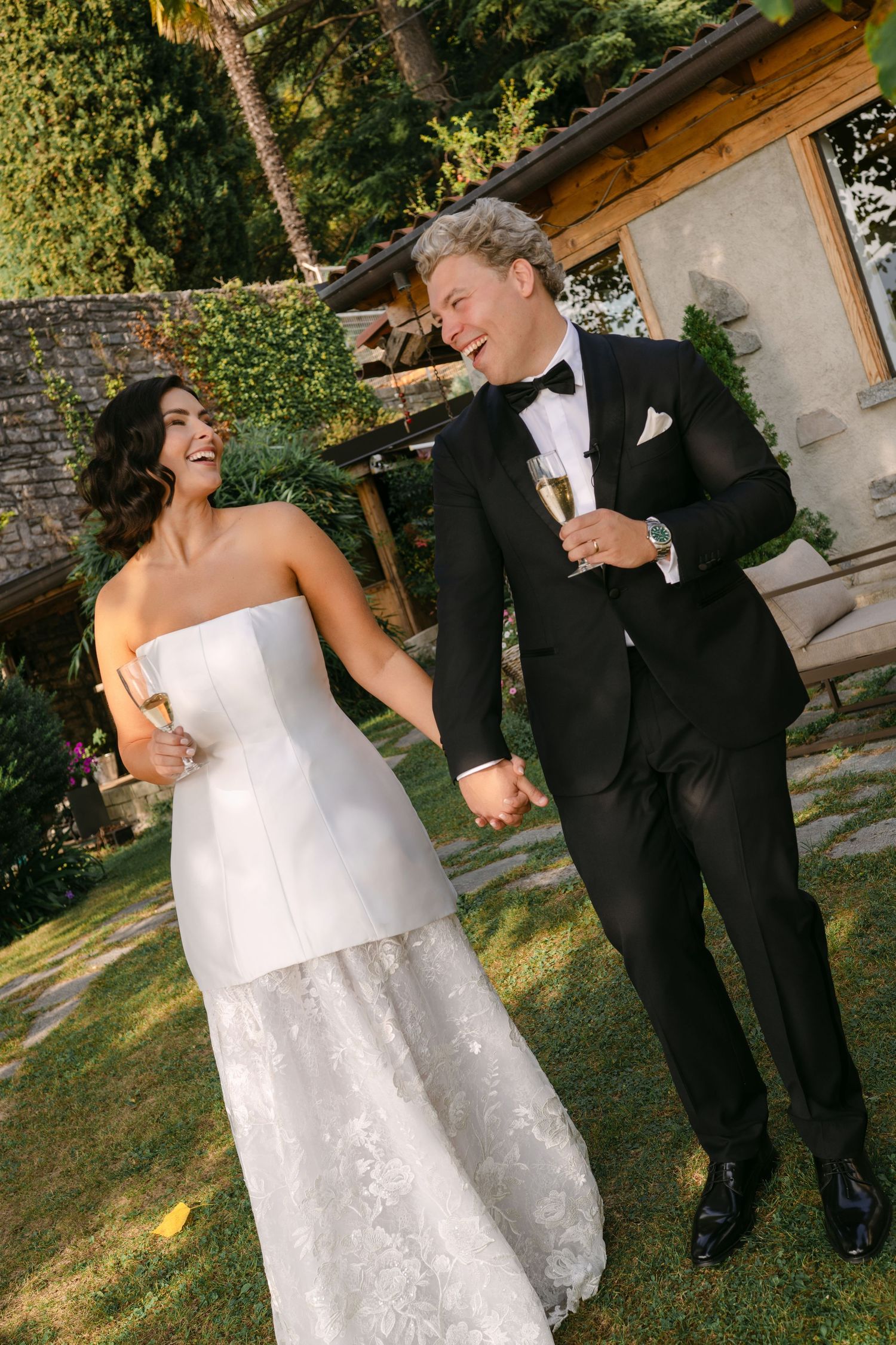 Laughing bride in a strapless white lace gown holds hands with a groom in a black tuxedo, both holding champagne glasses in a sunny garden.