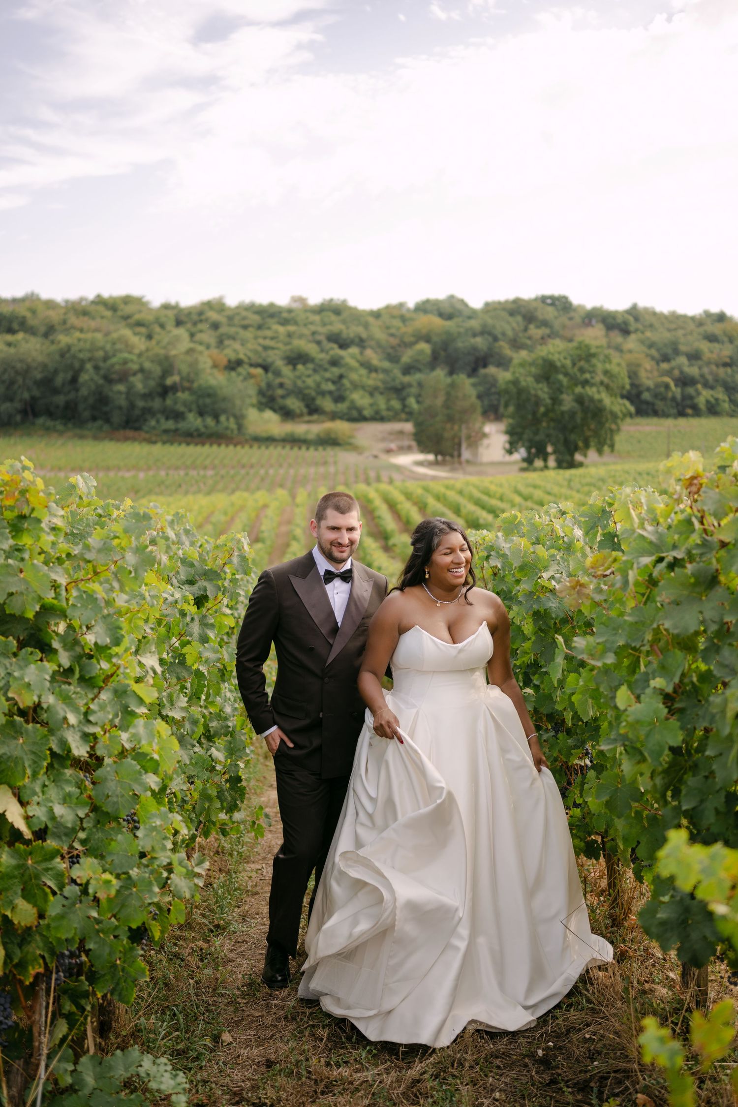 Bride and groom walk through lush vineyard rows in wedding attire, white gown flowing, with green hills in background.