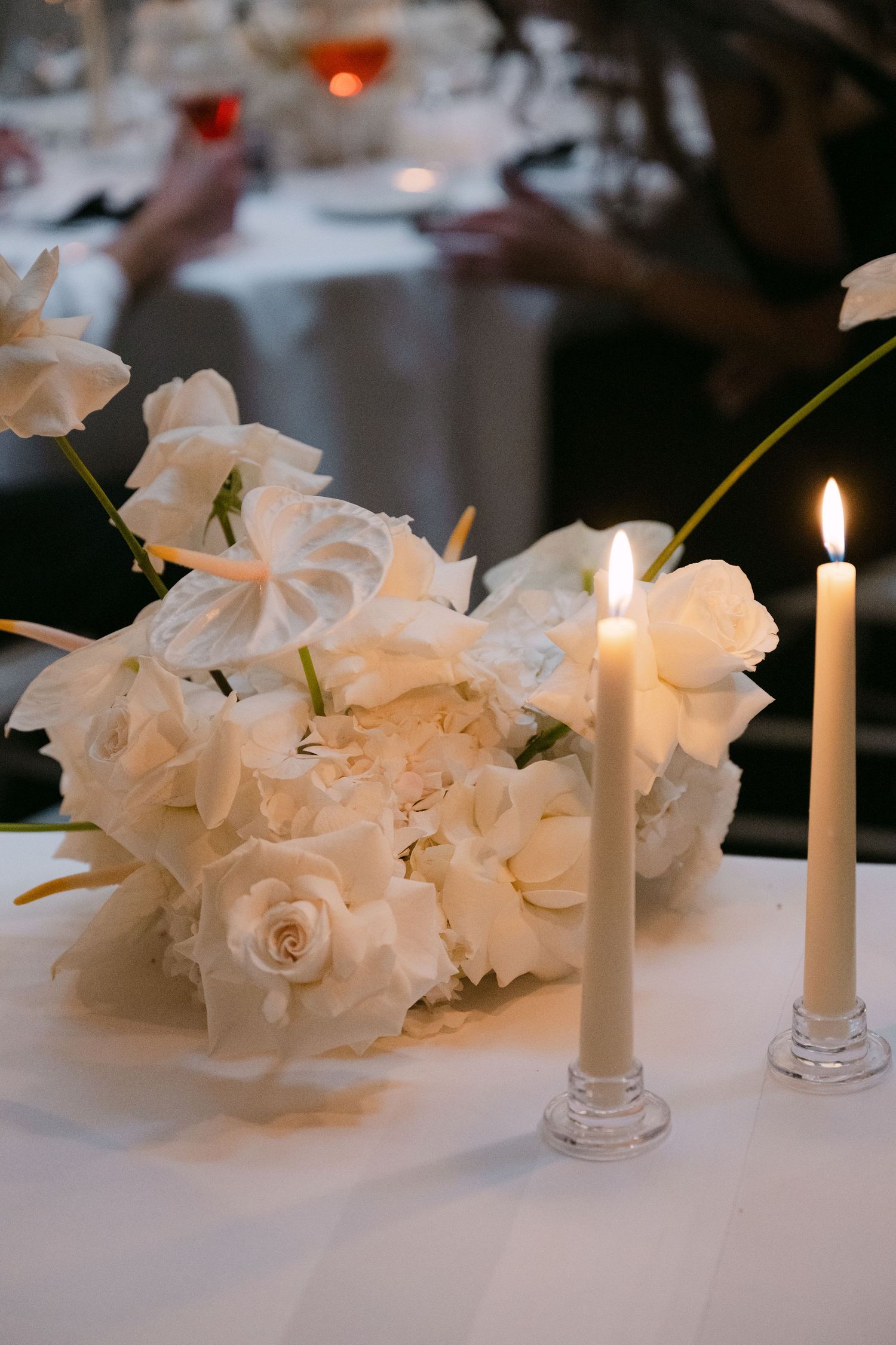 White floral centerpiece with lit taper candles on a wedding reception table.