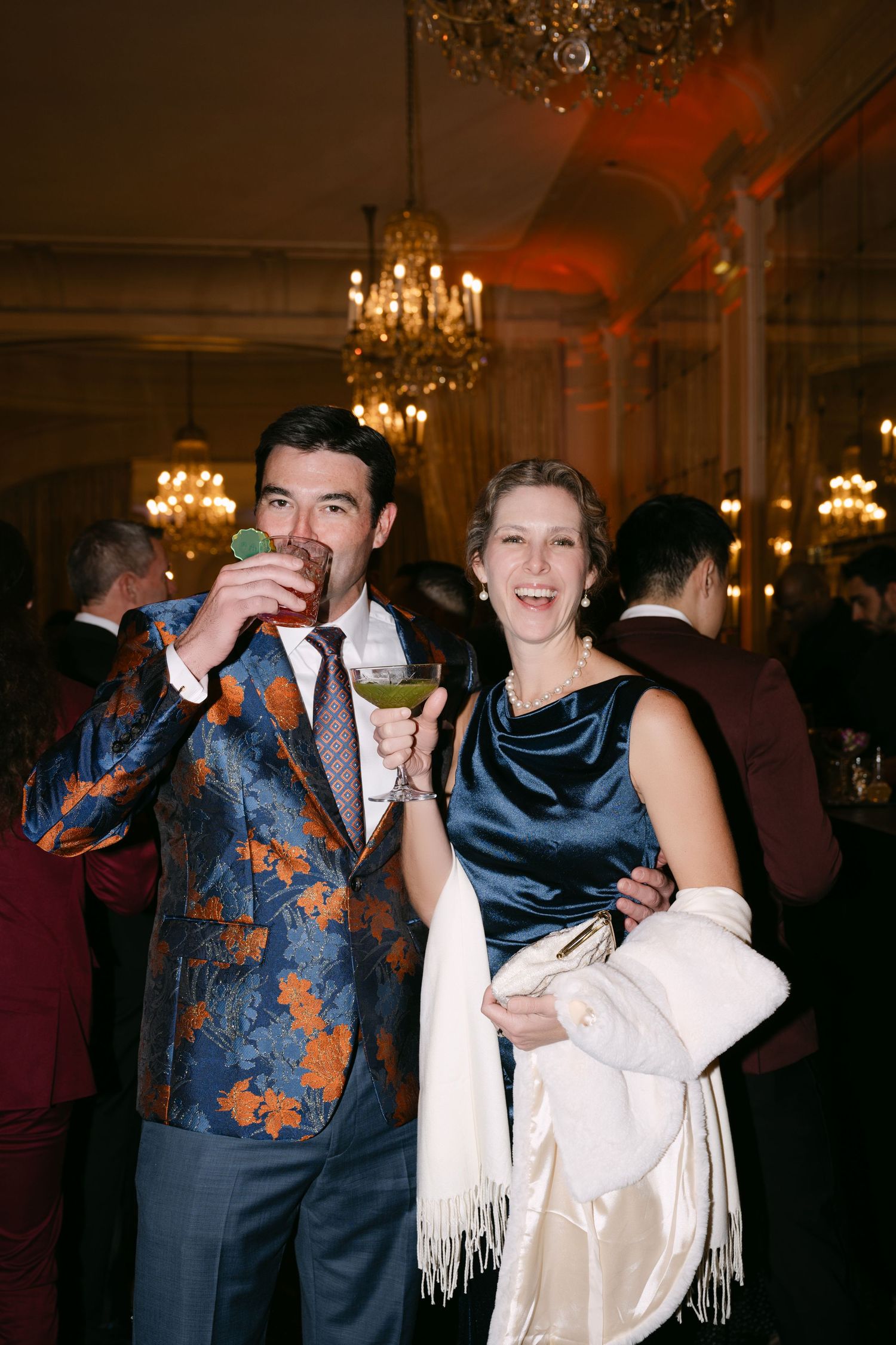 Stylish couple posing with cocktails at a formal wedding reception, man in floral blazer.