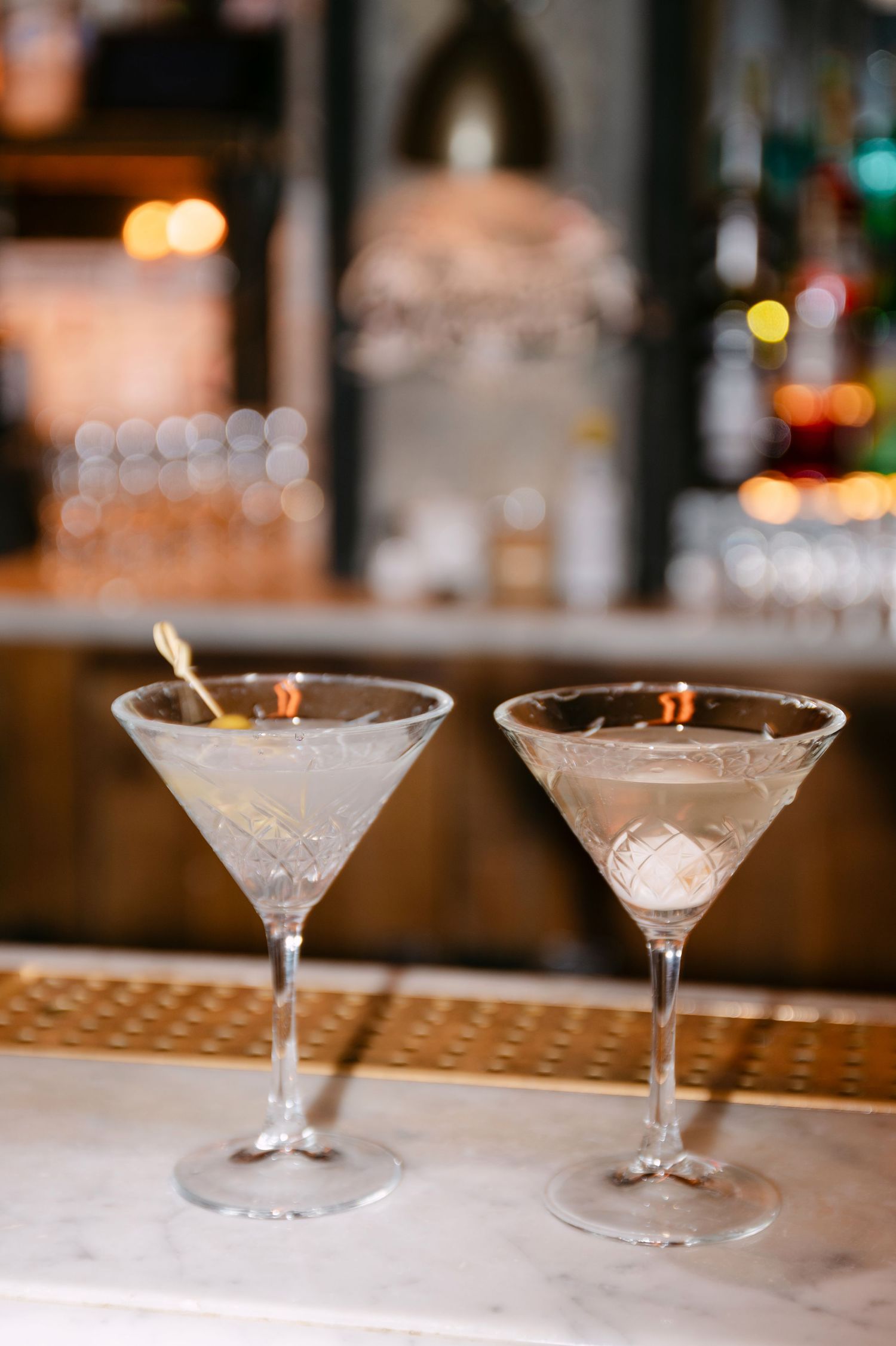 Two elegant crystal martini cocktails served on marble bar counter with bokeh lights.