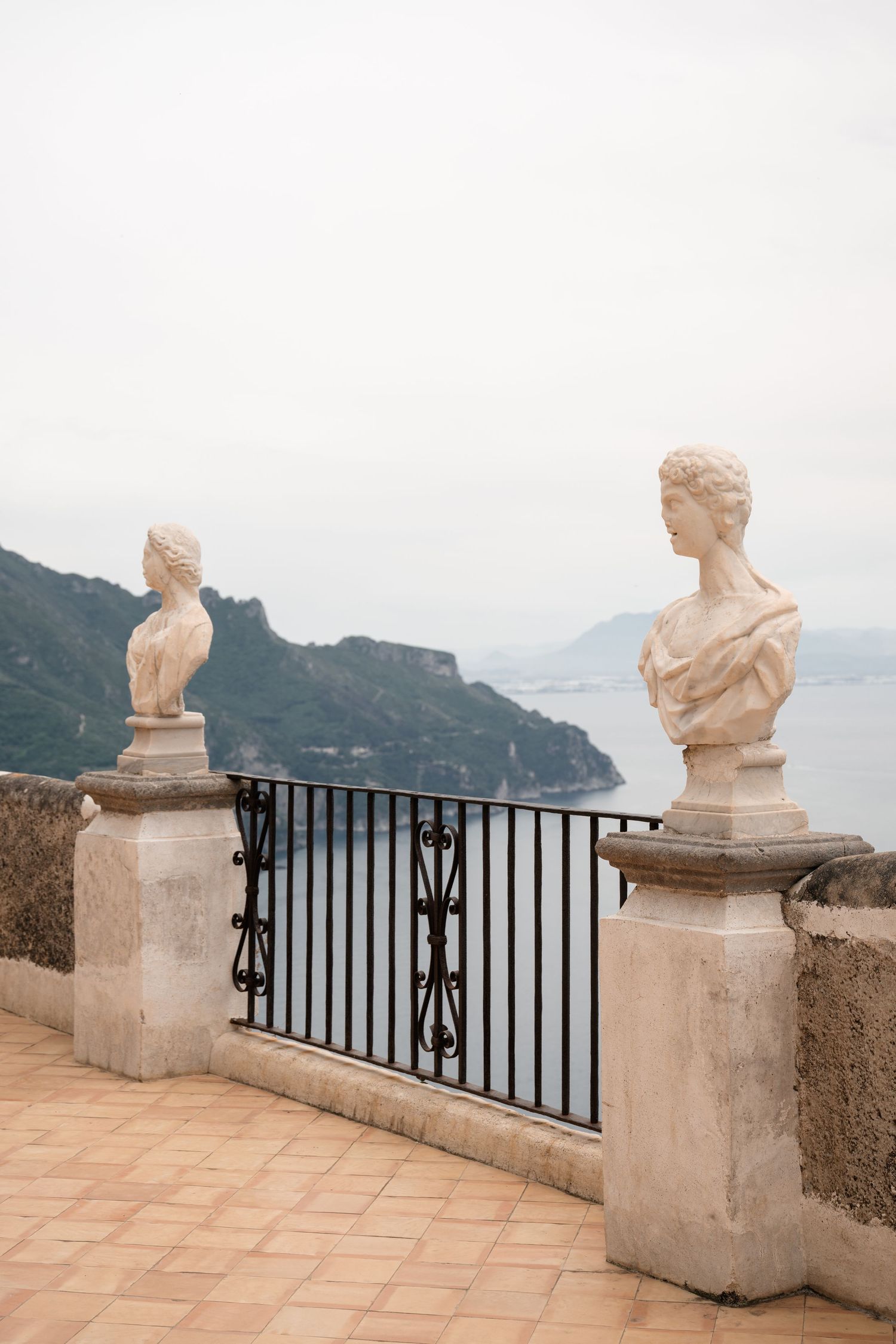 Two classical marble bust sculptures on terrace balustrade overlooking Amalfi Coast and sea.