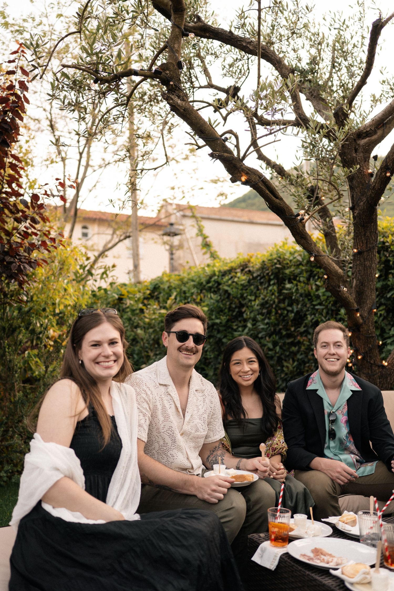 Four smiling wedding guests enjoy drinks and food together at an outdoor garden party in Italy.