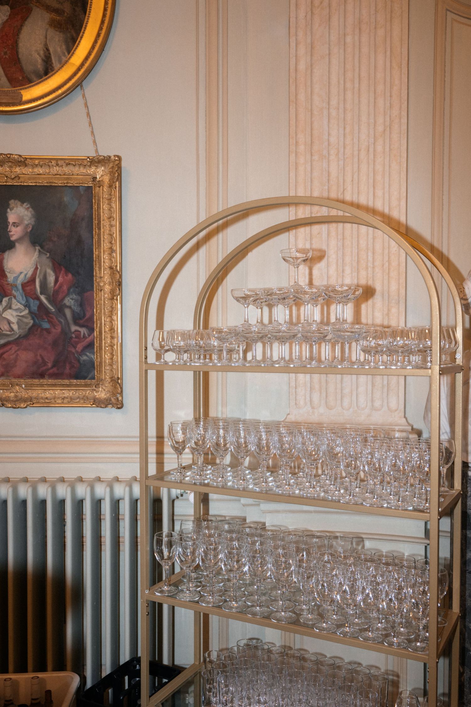 Gold tiered bar cart displaying crystal champagne coupes and wine glasses in an ornate French interior.