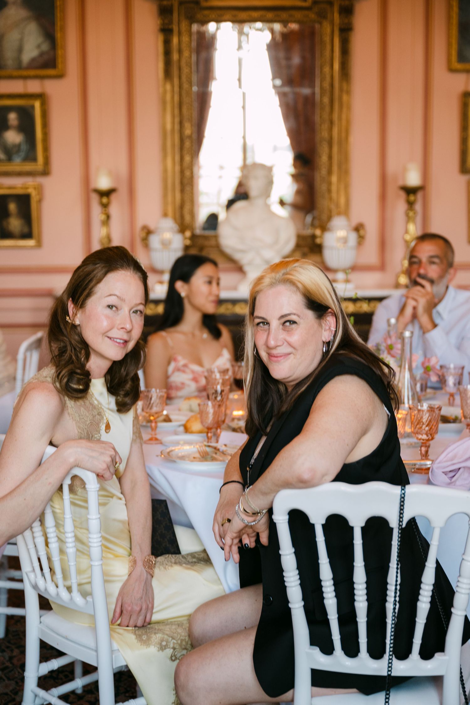 Two smiling women pose together at a wedding reception in an elegant pink French chateau dining room.
