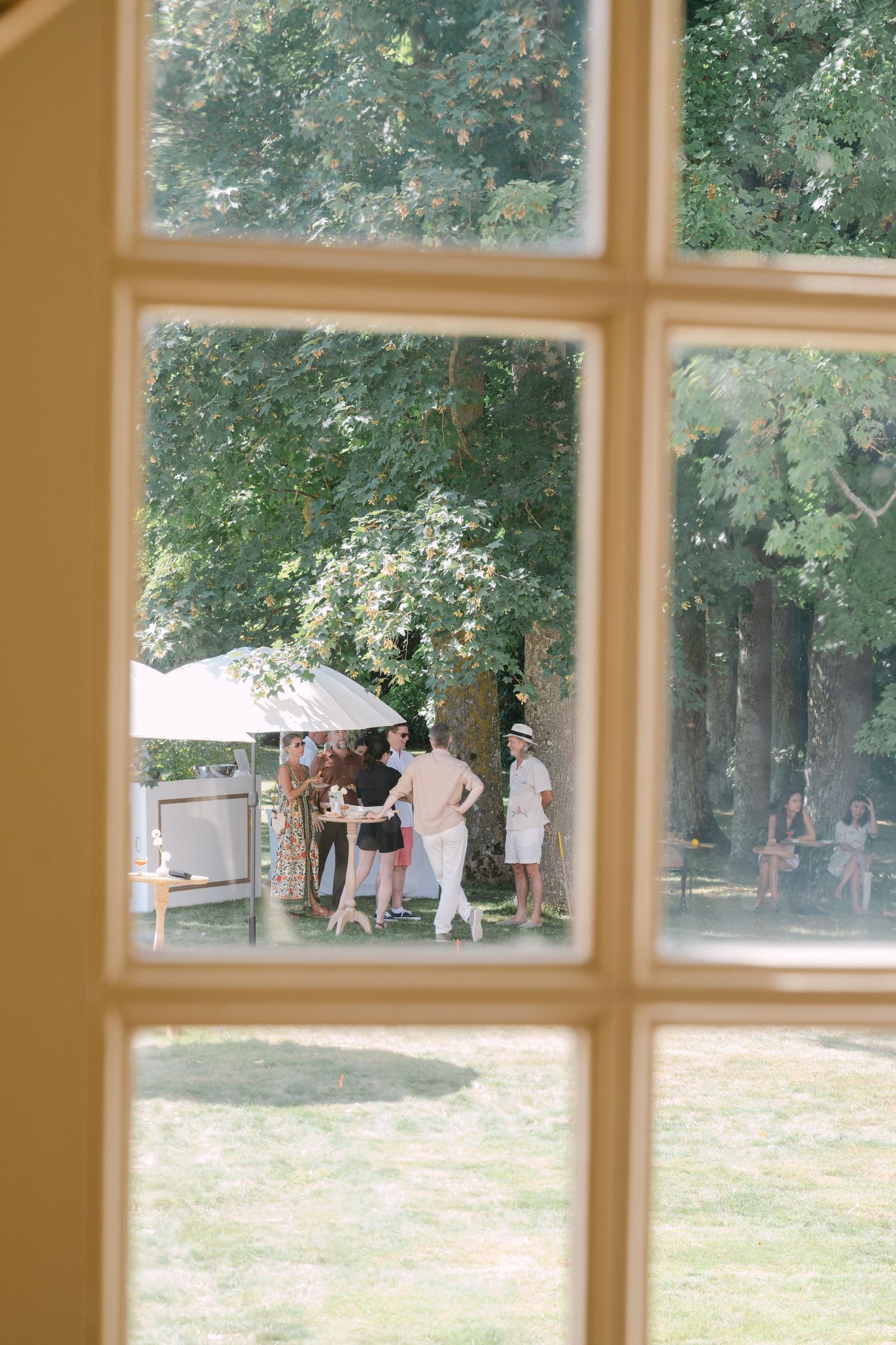 View through a French window of wedding guests mingling under trees at an outdoor garden reception in summer.