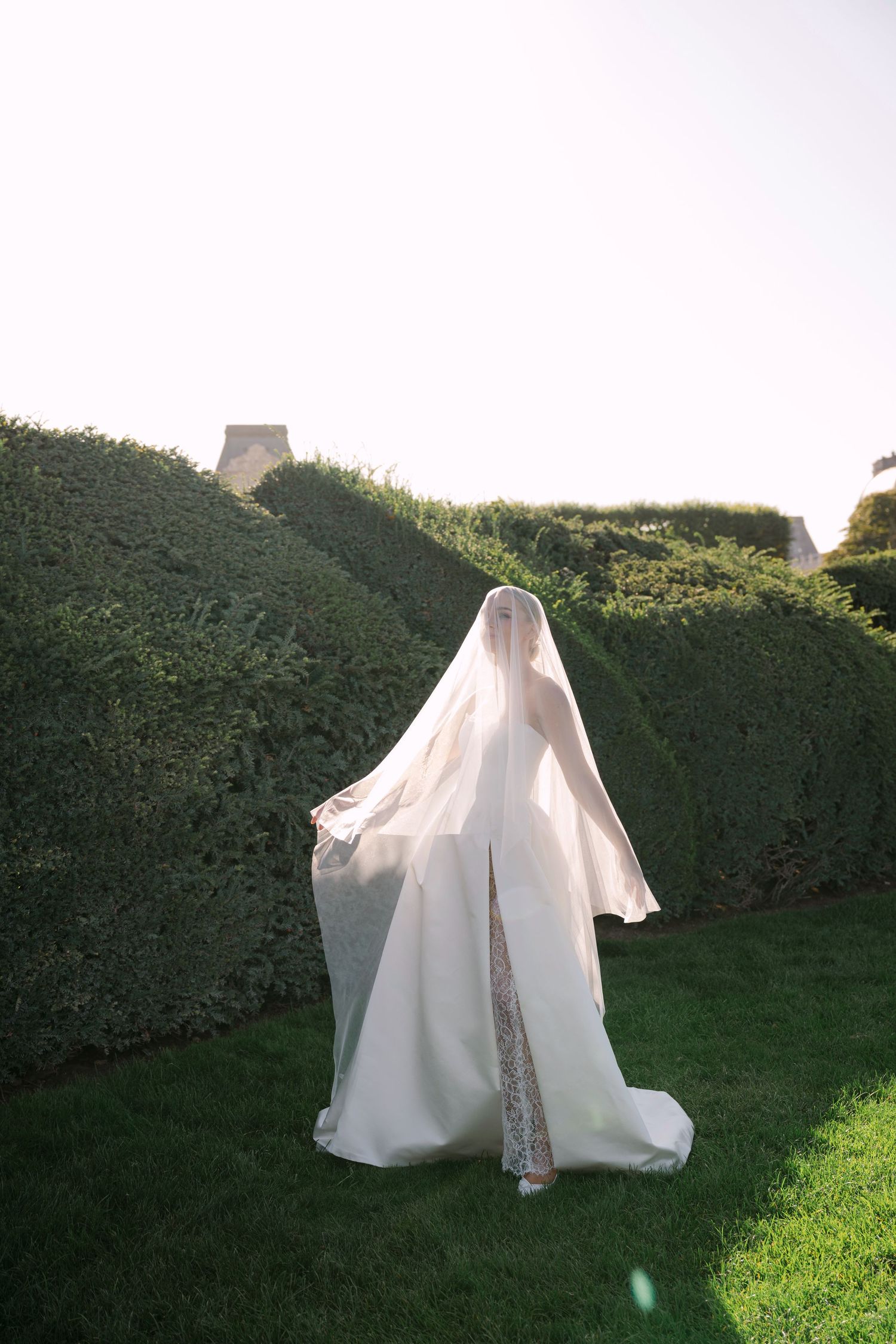 Bride in flowing white gown and cathedral veil walks through manicured green hedges in a garden.