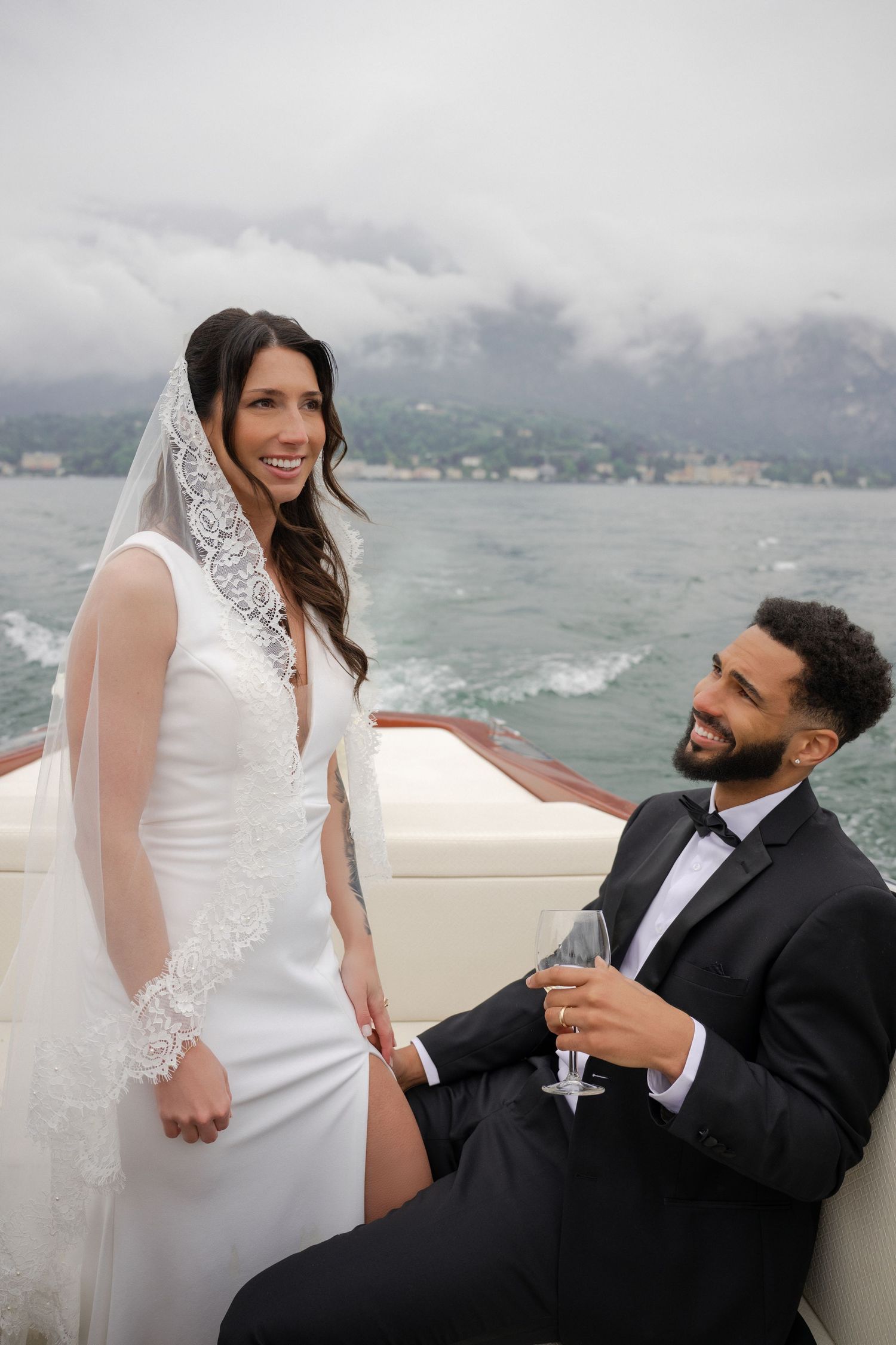 Bride and groom celebrate on luxury boat on Lake Como, groom holding champagne glass on cloudy day.