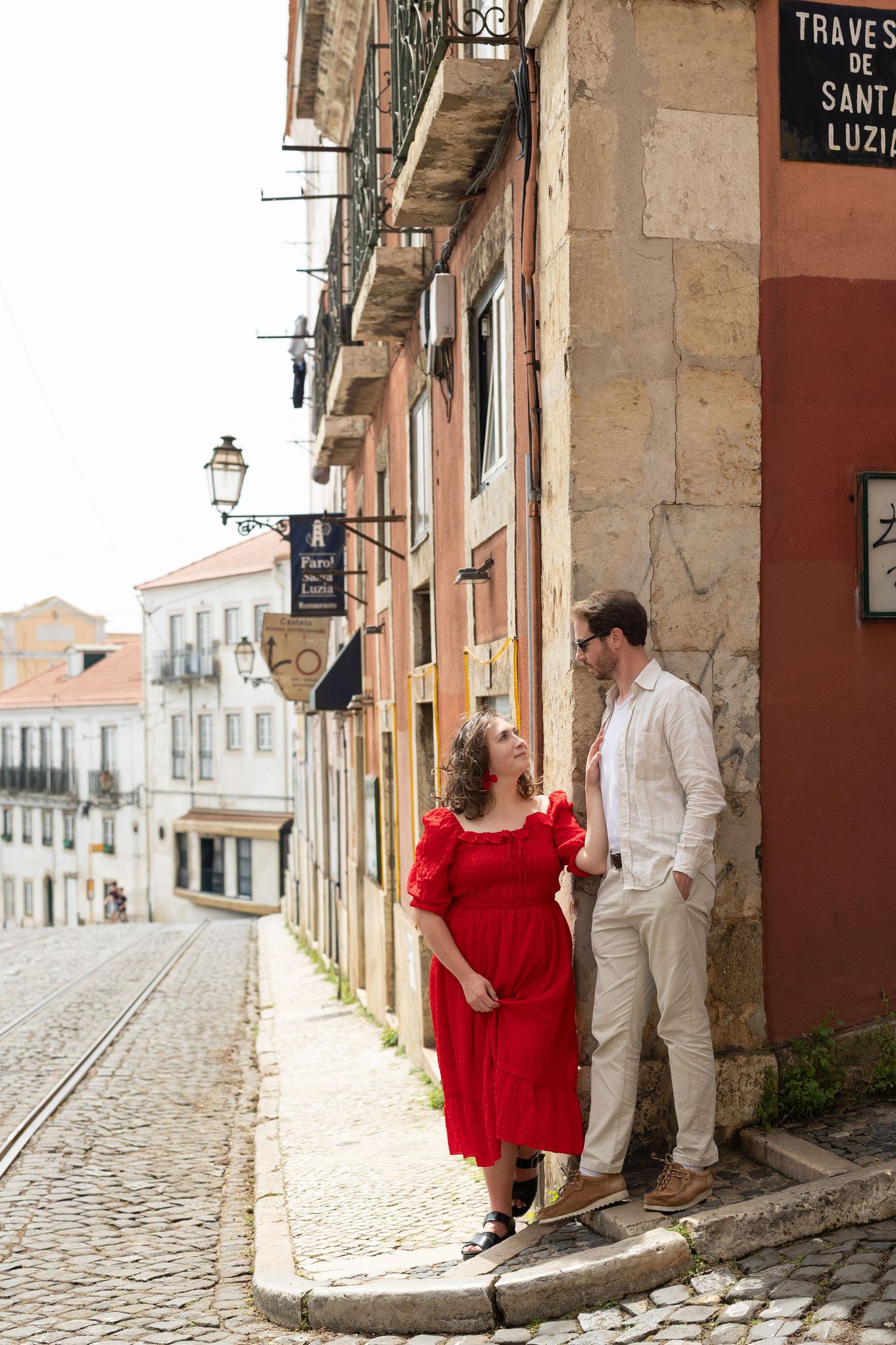 Couple posing on a cobblestone street in Porto, woman in red dress, man in white shirt, colorful buildings backdrop.