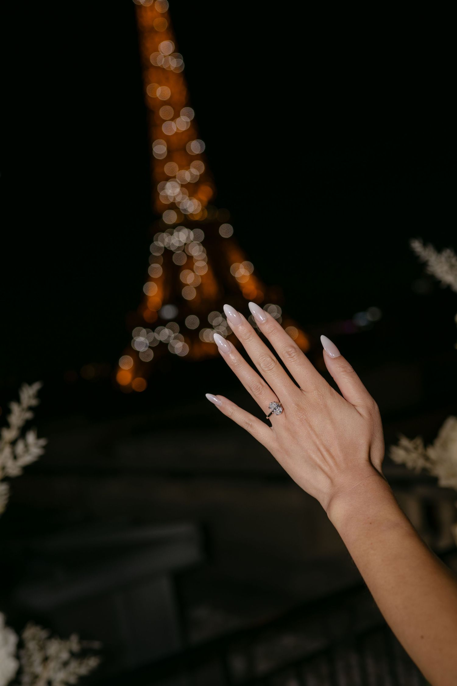 Woman's hand with a diamond engagement ring raised in front of the glowing Eiffel Tower at night in Paris.