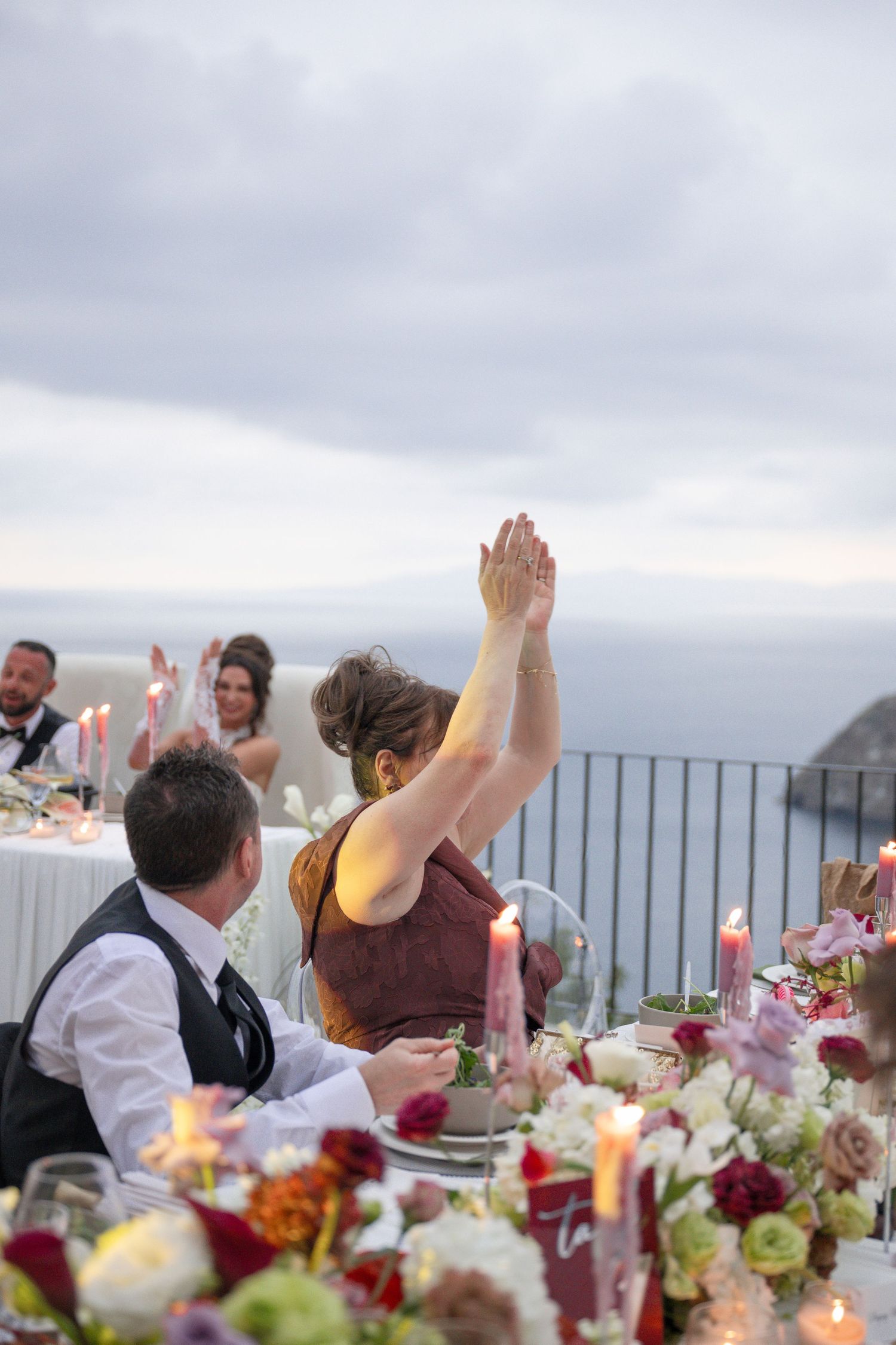Wedding guest raising hands at candlelit outdoor reception table with ocean view at dusk.