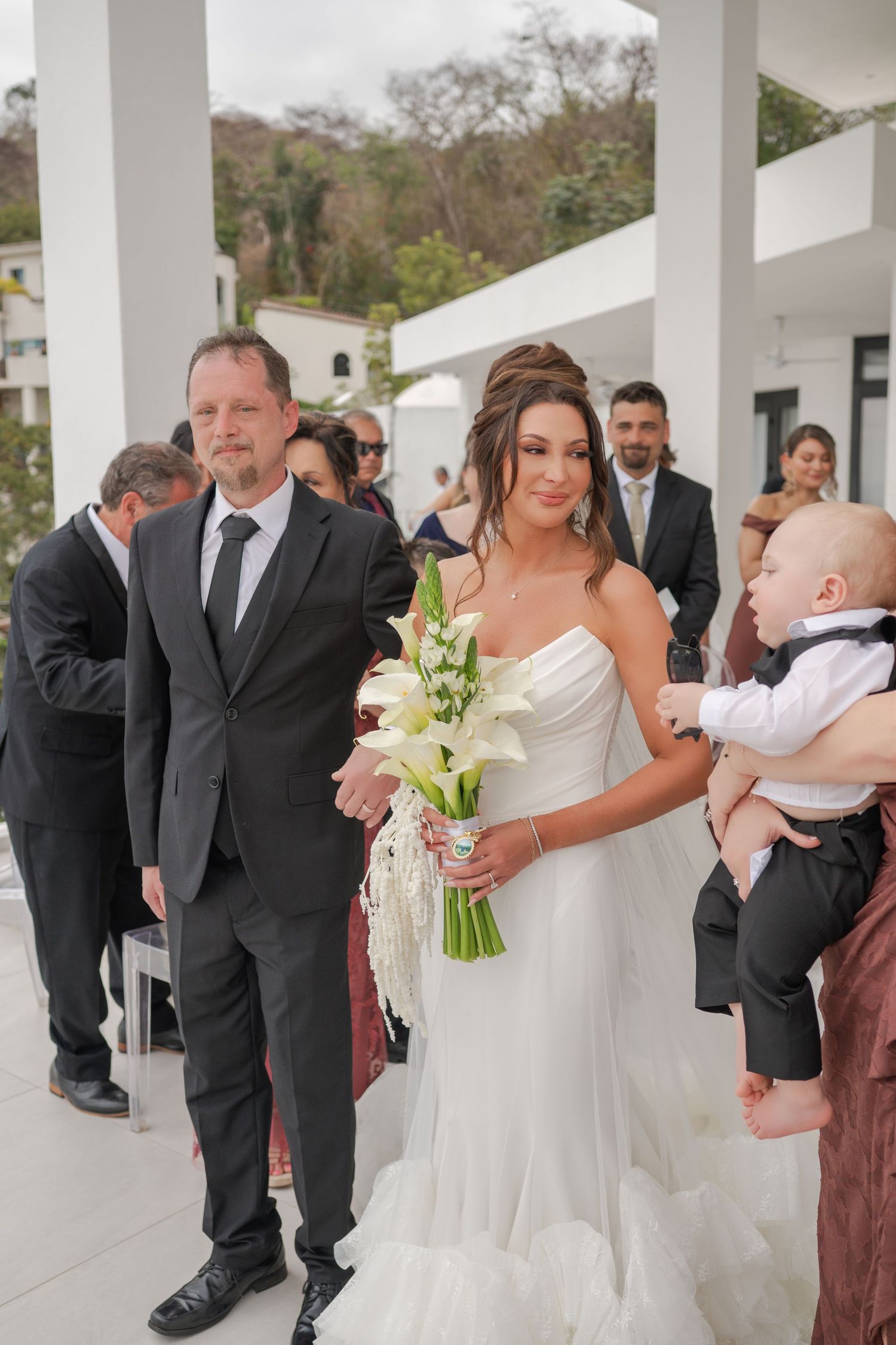 Bride in white strapless gown holding calla lily bouquet walks down aisle with man in gray suit at outdoor wedding.
