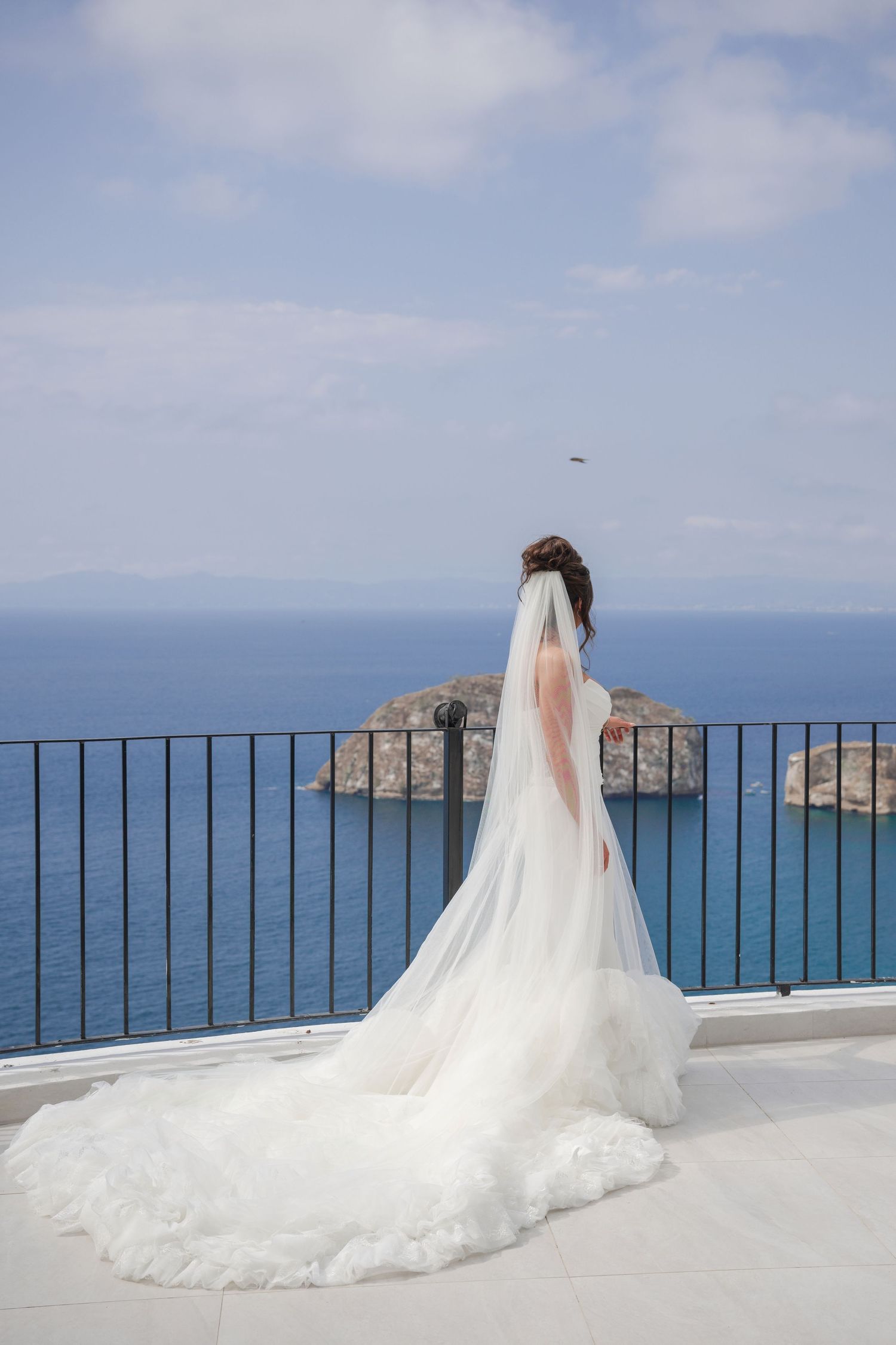 Bride in a white gown and cathedral veil gazes at rocky ocean islands from a balcony.