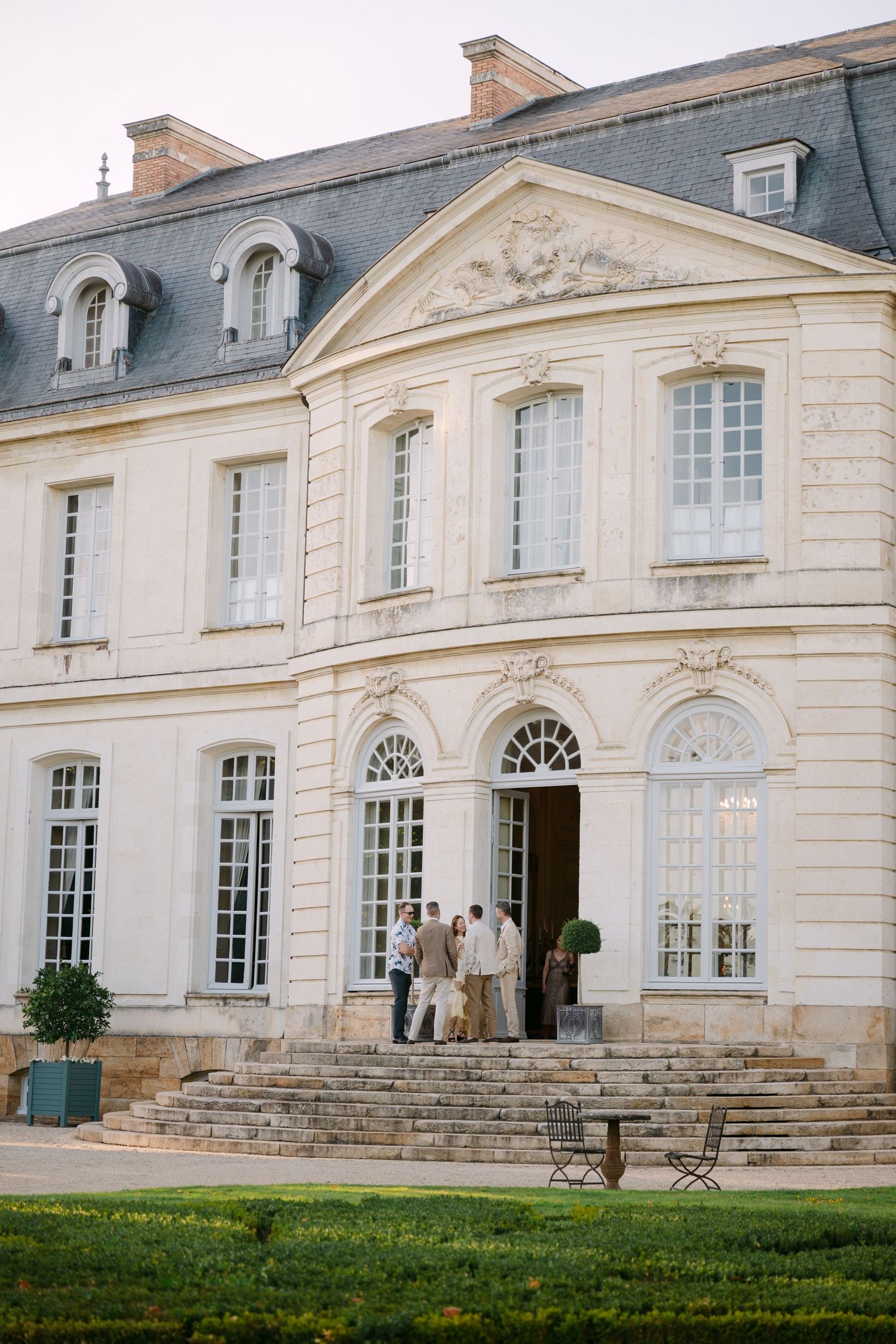 Grand French neoclassical chateau exterior with white stone facade, arched windows, and manicured grounds at dusk.