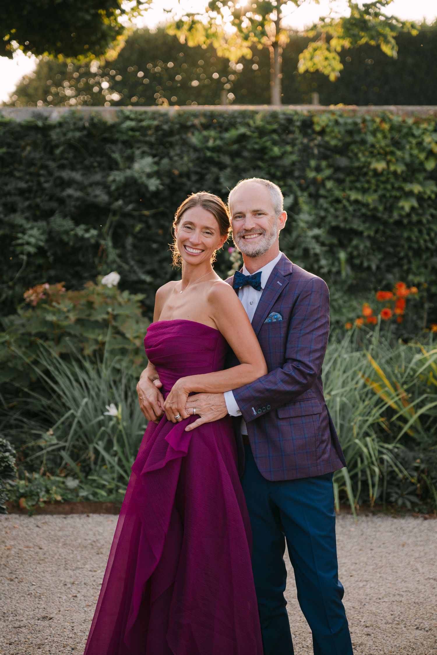 Couple posing at golden hour in garden, woman in purple gown and man in plaid blazer.