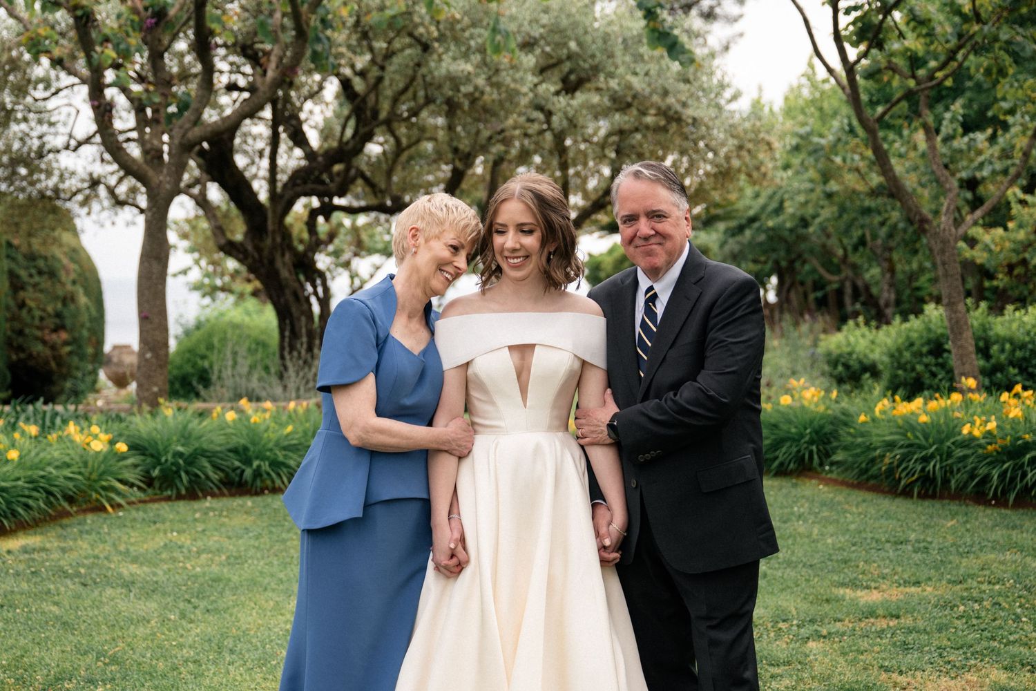 Bride in elegant off-shoulder gown poses with parents in lush garden setting.