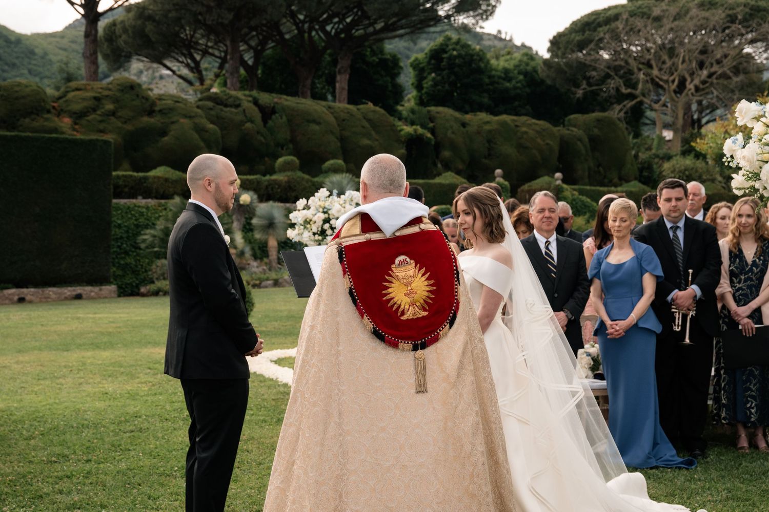 Bride and groom exchange vows with officiant during elegant outdoor wedding ceremony in lush garden setting.