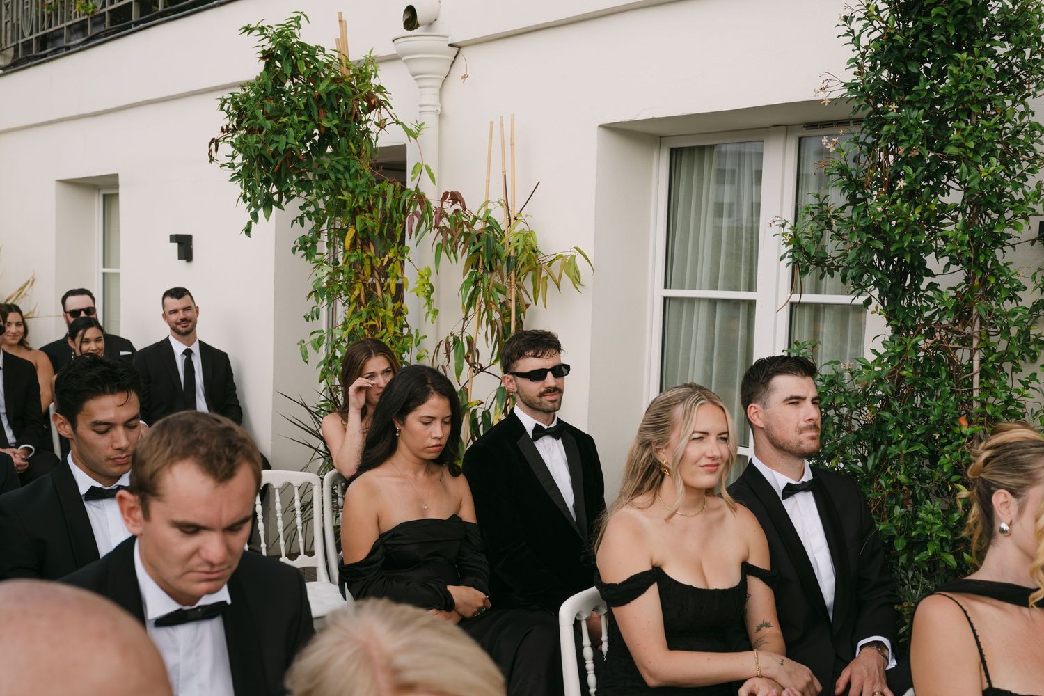 Wedding guests dressed in black formal attire seated outdoors at elegant wedding ceremony.