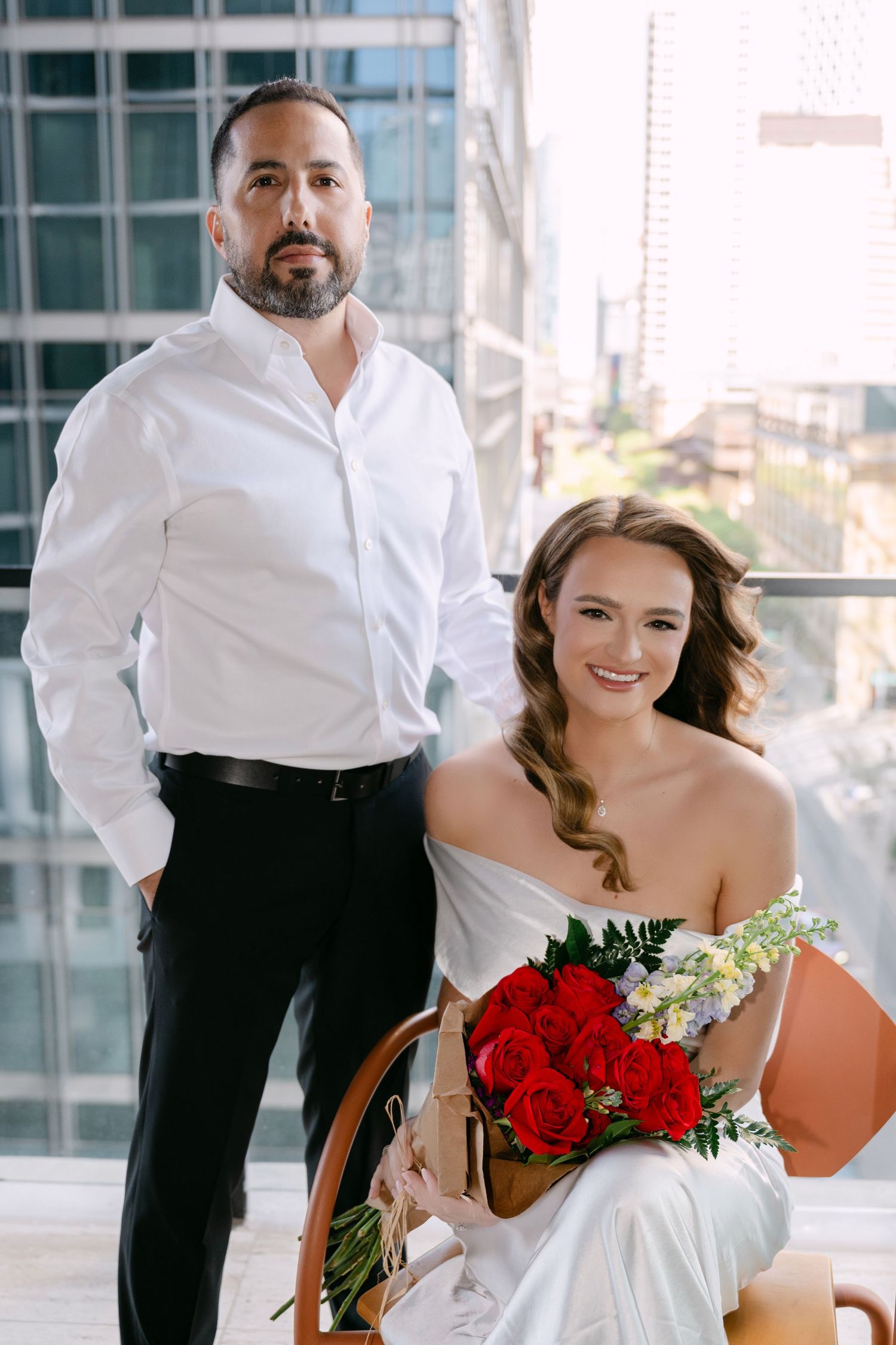 Bride and groom posing on a city high-rise balcony, bride seated holding red roses bouquet in white gown.