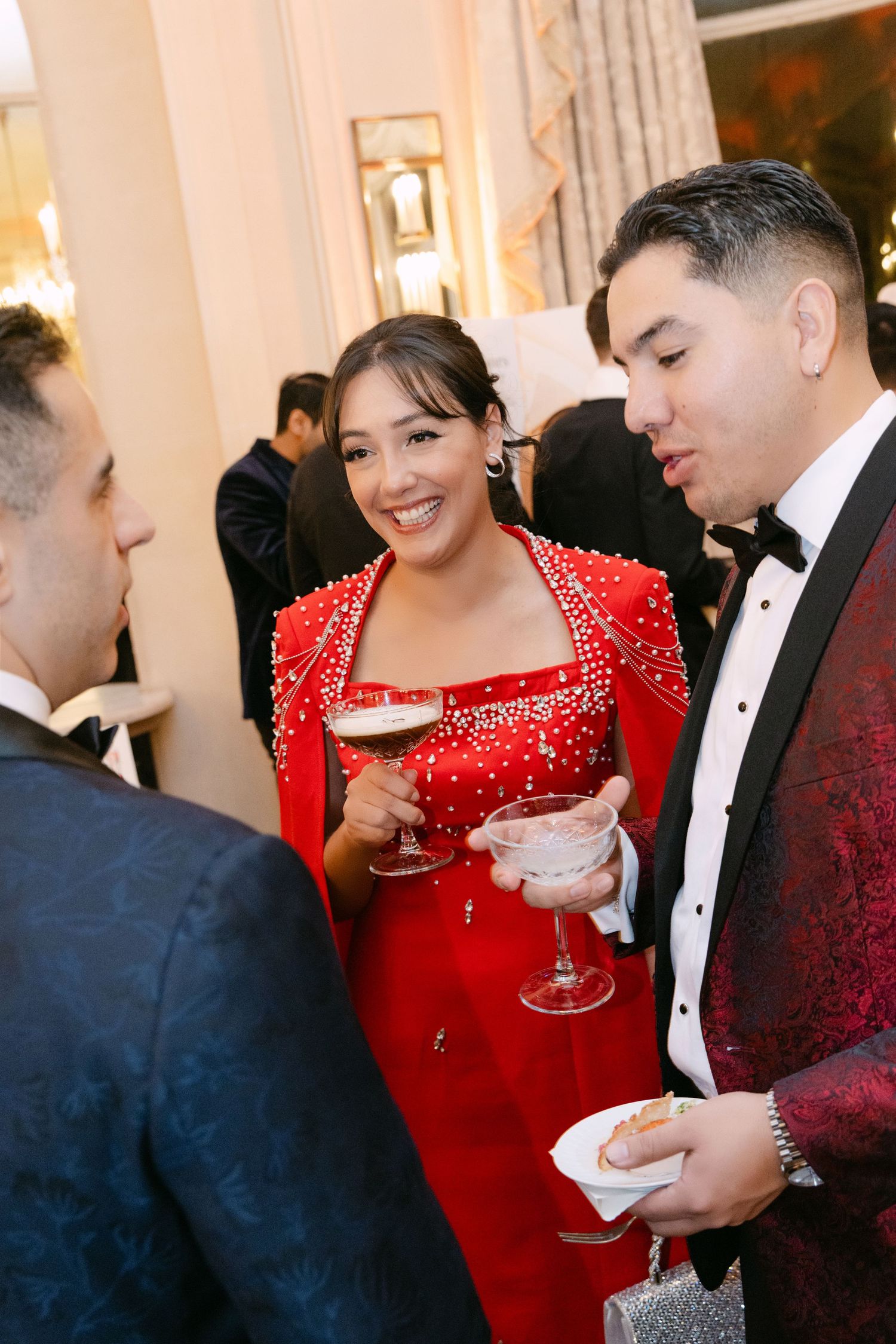 A woman in a red beaded gown smiles while holding a martini glass, chatting with guests at a formal wedding reception.
