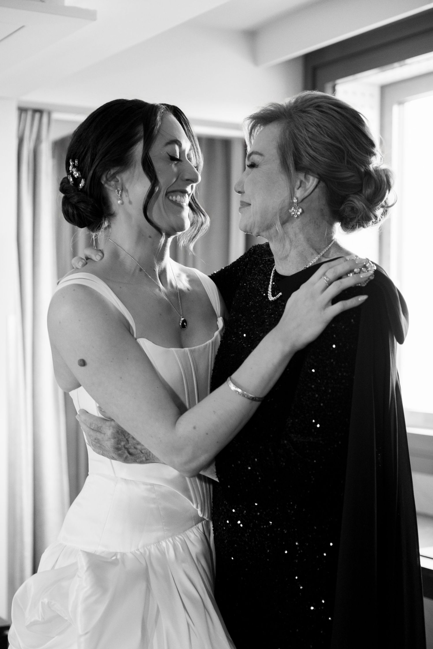 Black and white photo of smiling bride in white gown embracing older woman in black sparkly dress.