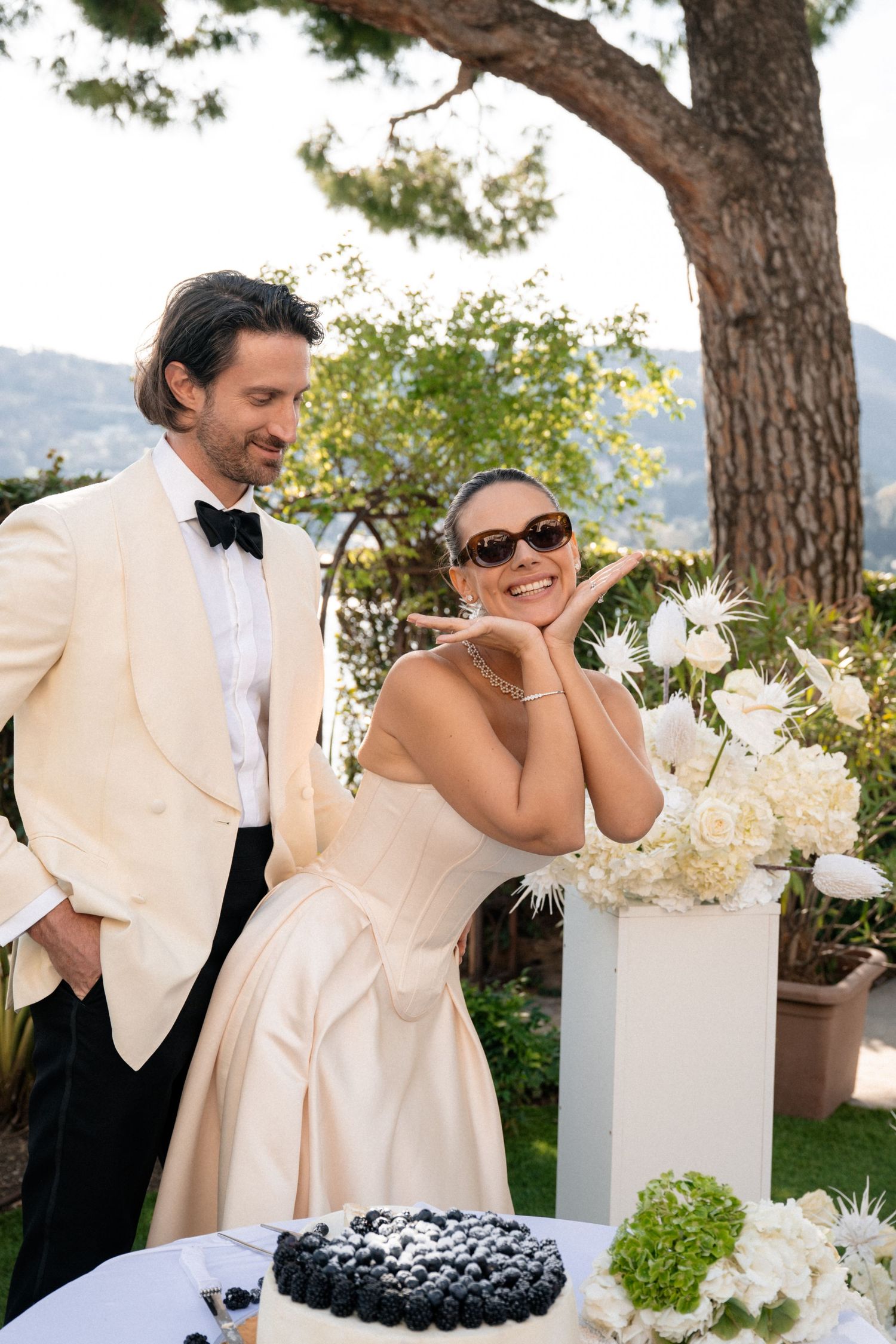 Smiling bride in strapless gown poses with groom in ivory tuxedo beside wedding cake and white florals.