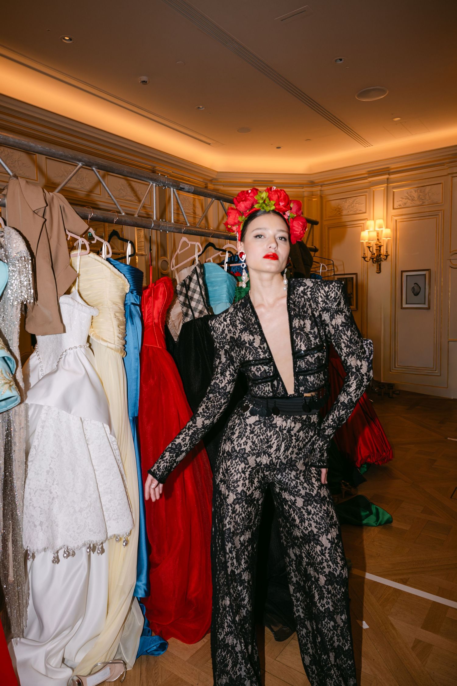 Model in black lace jumpsuit with red floral crown stands beside garment rack backstage.