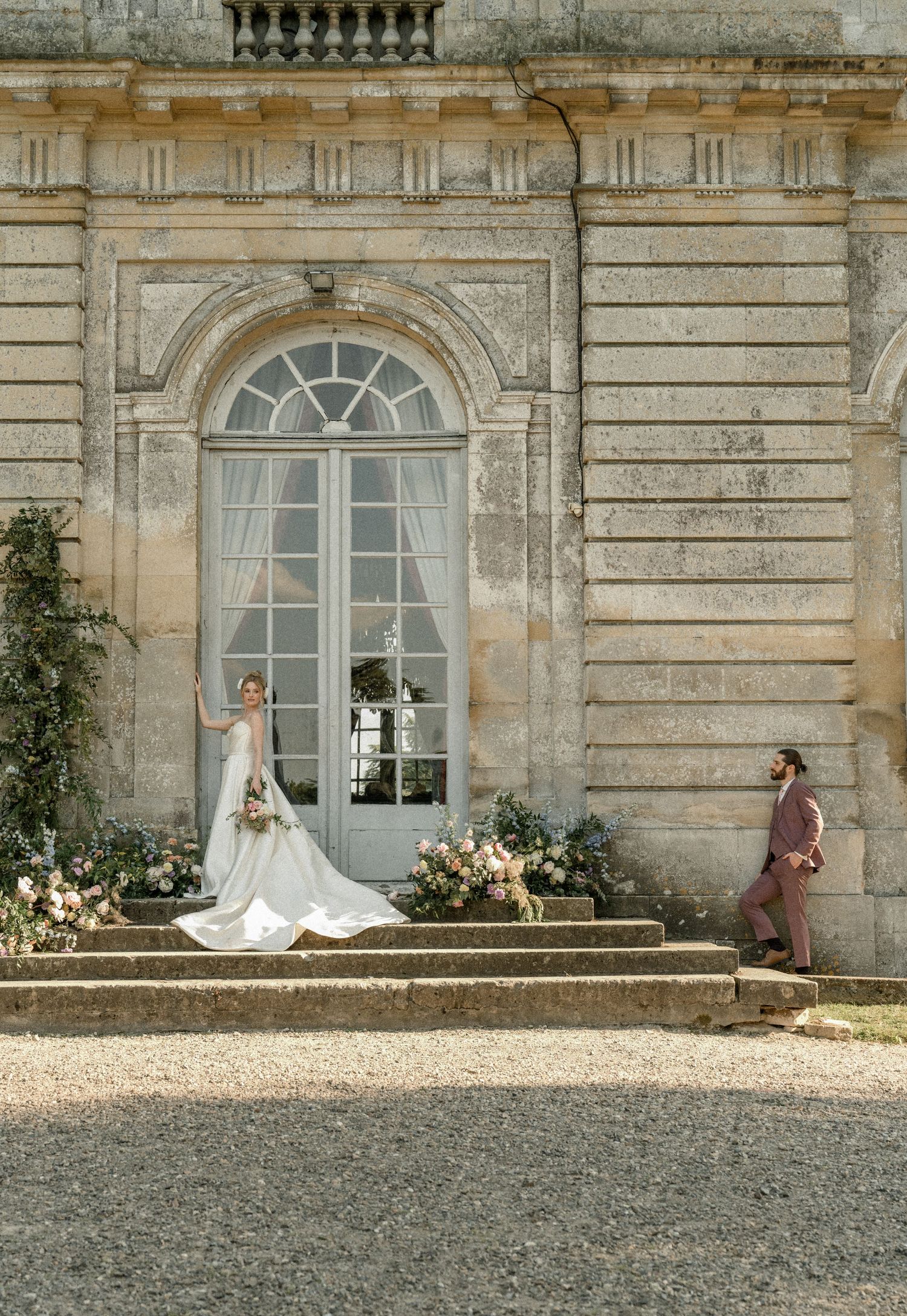 A bride poses dramatically on stone steps while someone stands nearby at a classical building entrance.