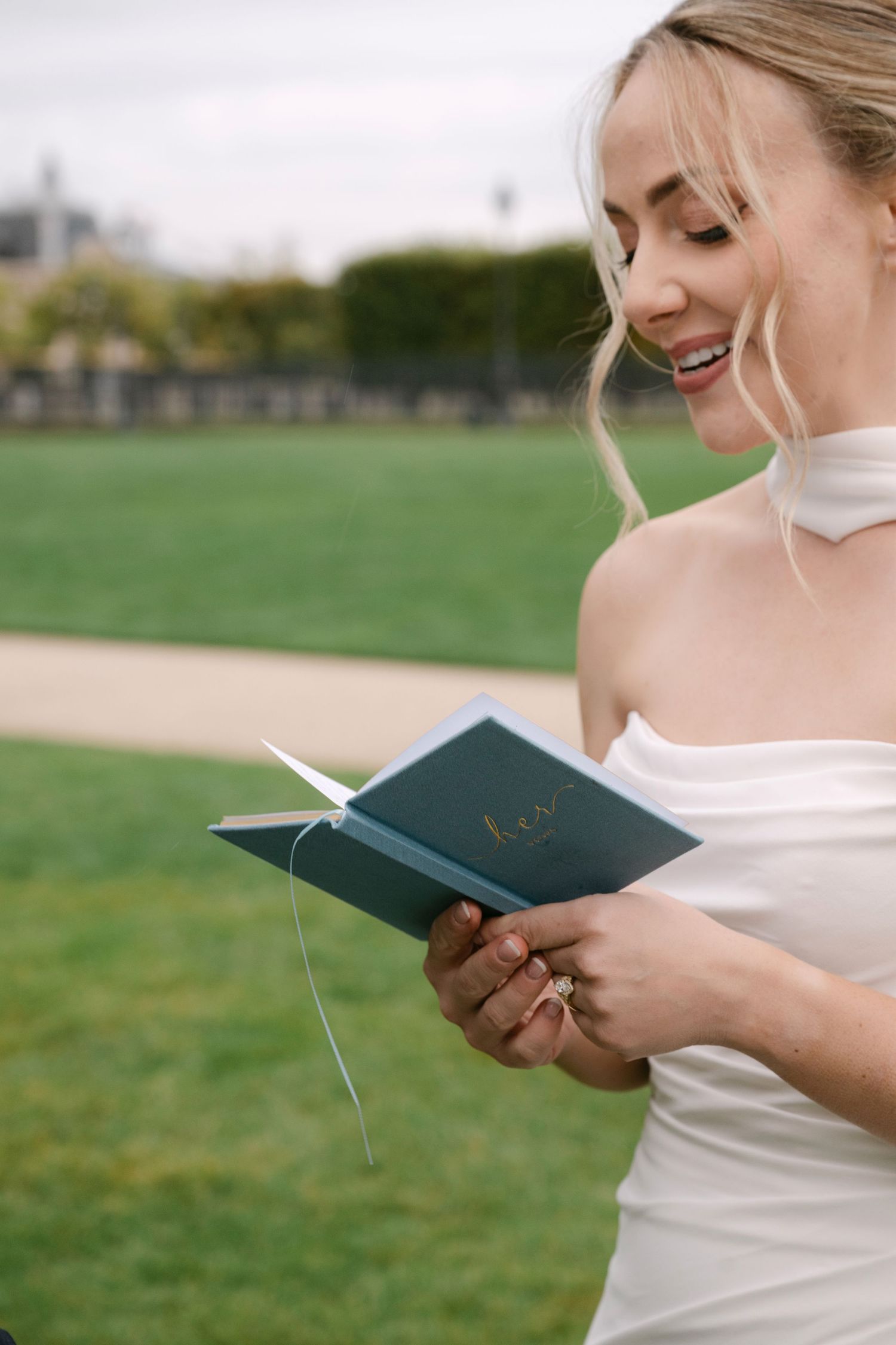 A person in a white wedding dress reads from a small book during an outdoor ceremony.