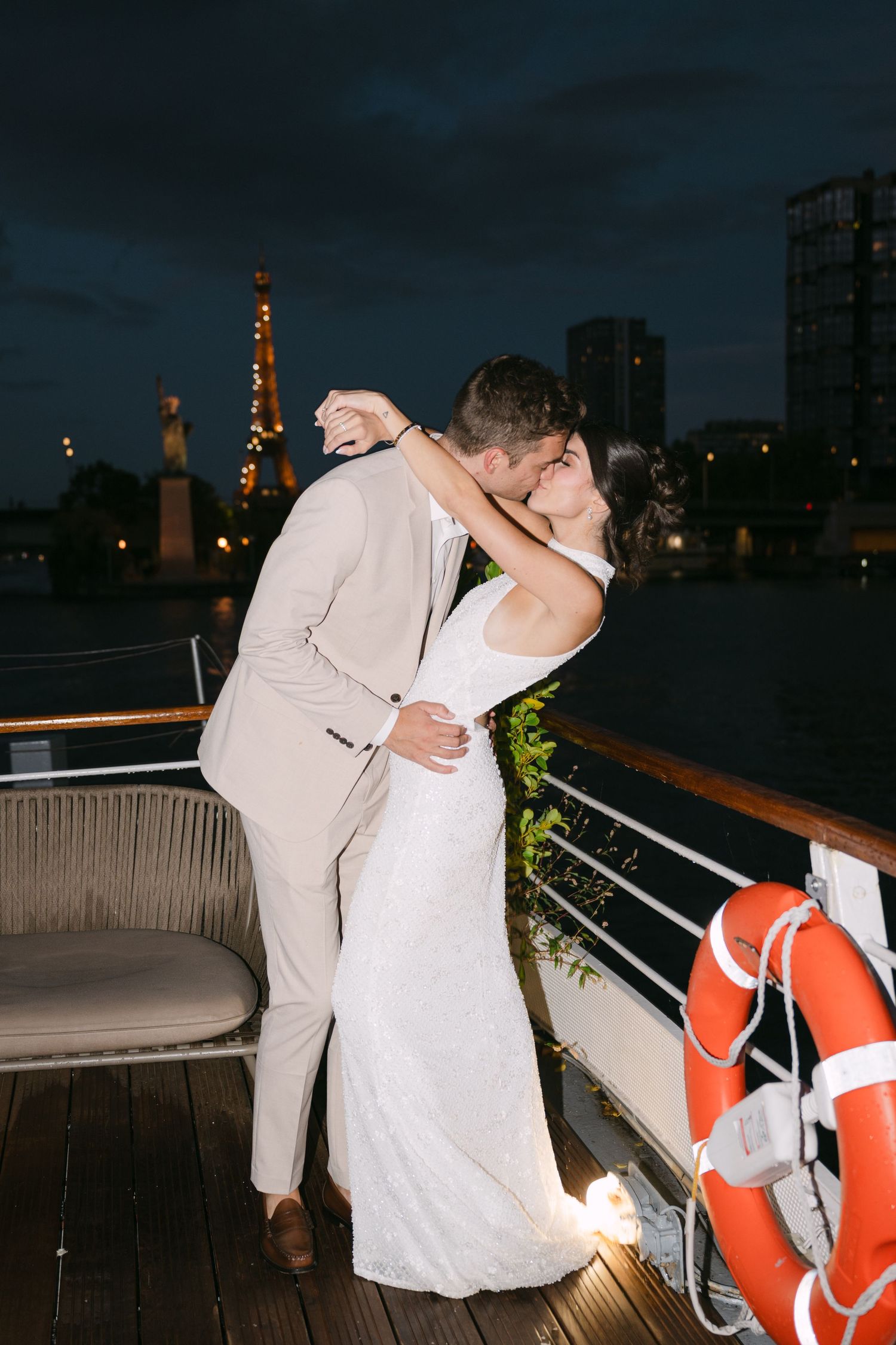 Newlywed couple sharing romantic kisses on a nighttime deck with orange life preservers and city lights in background.
