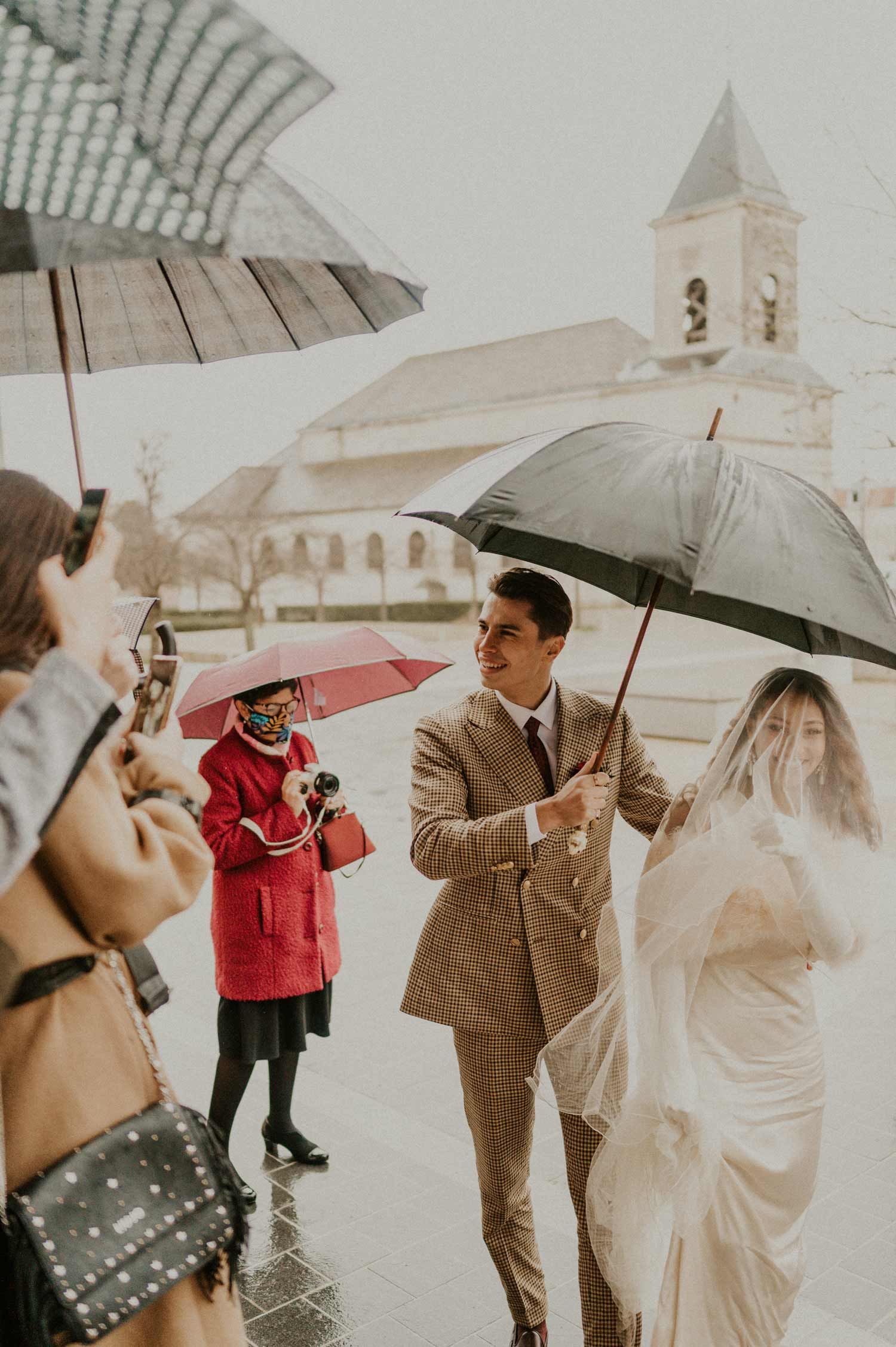 Wedding party holding umbrellas outside a church on a rainy day.