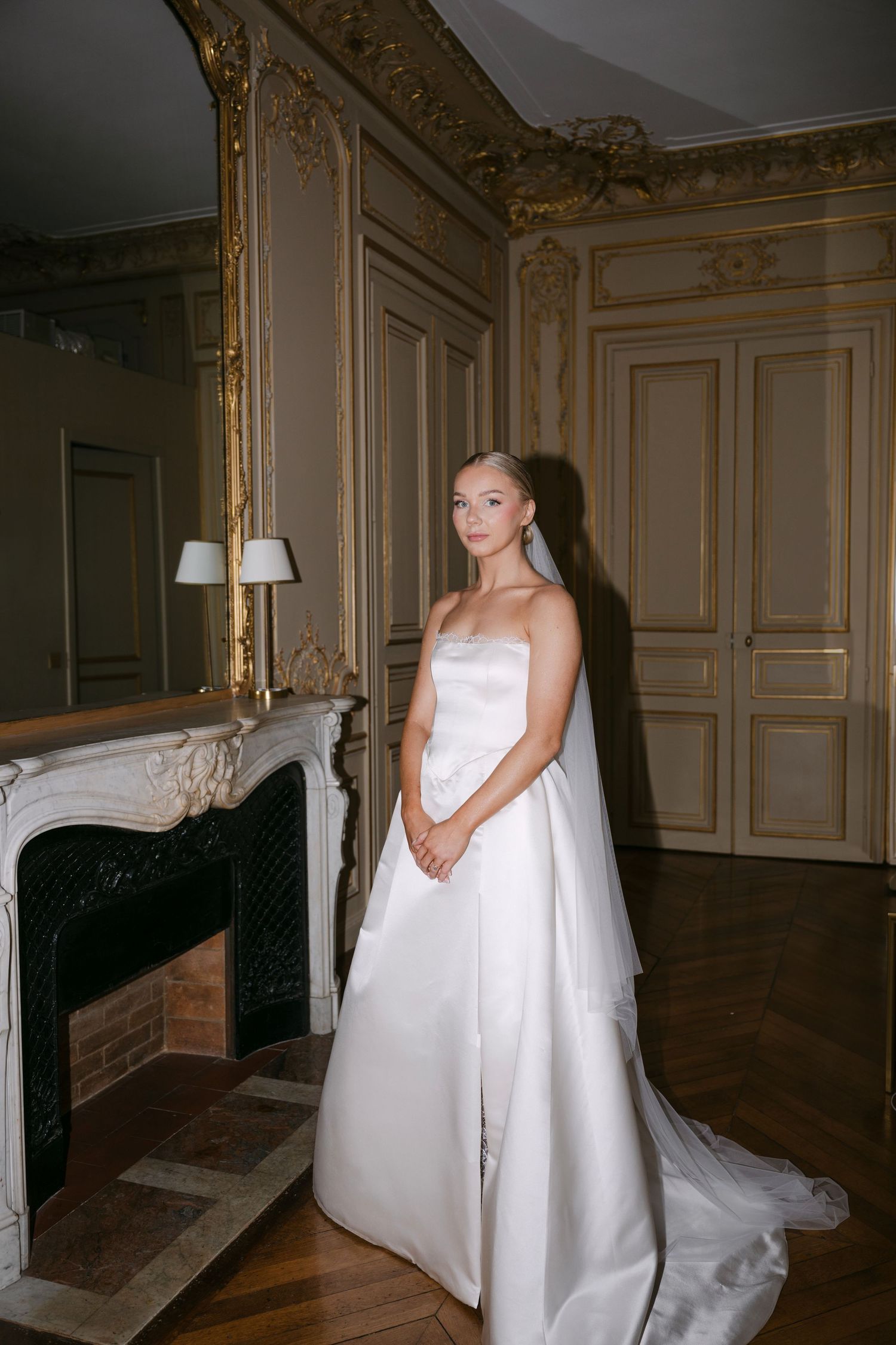 Bride in strapless white satin gown with veil poses by ornate fireplace in elegant Parisian room.
