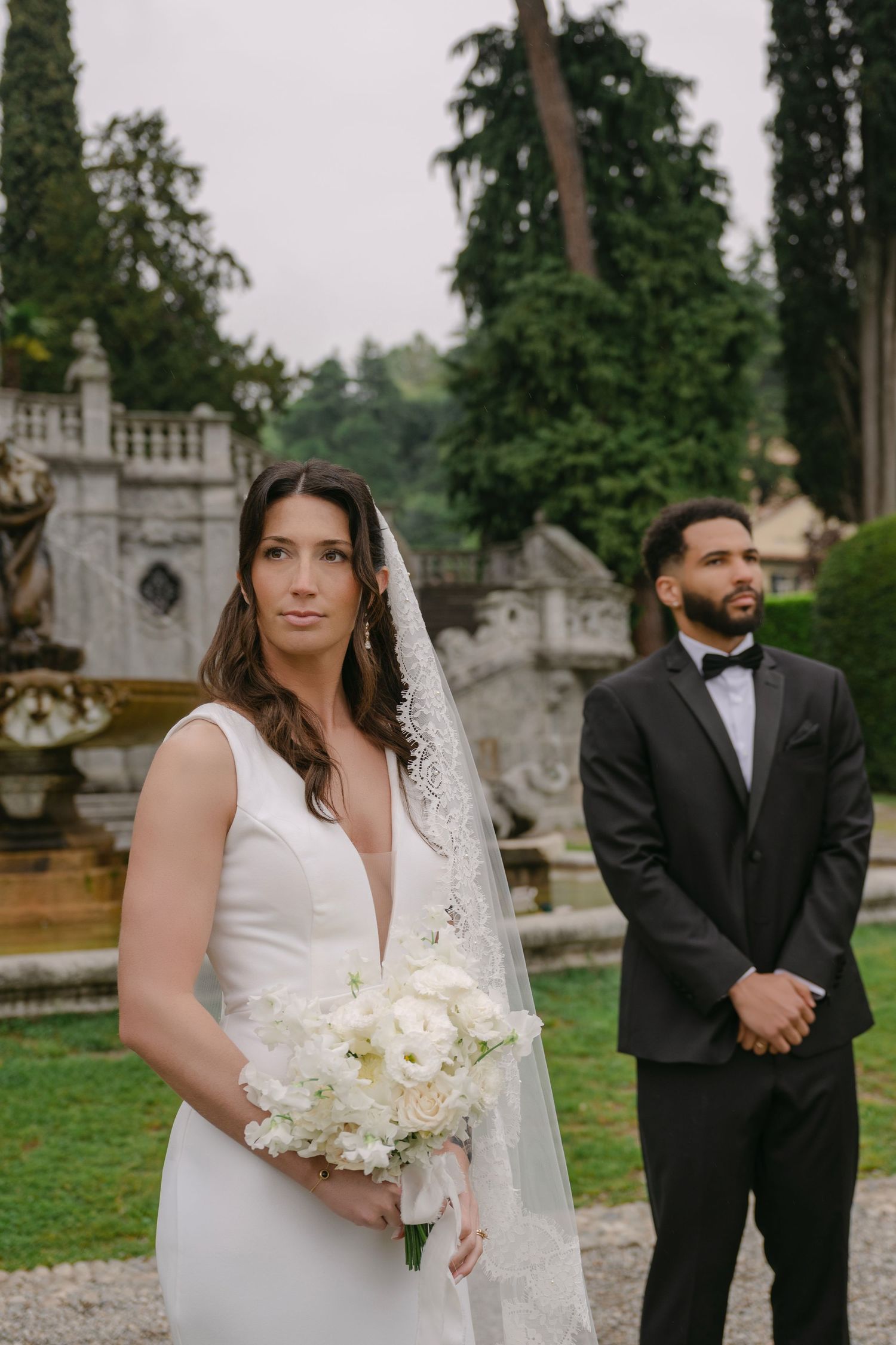 Bride in deep V-neck gown holding white bouquet with groom in black tuxedo in garden setting.