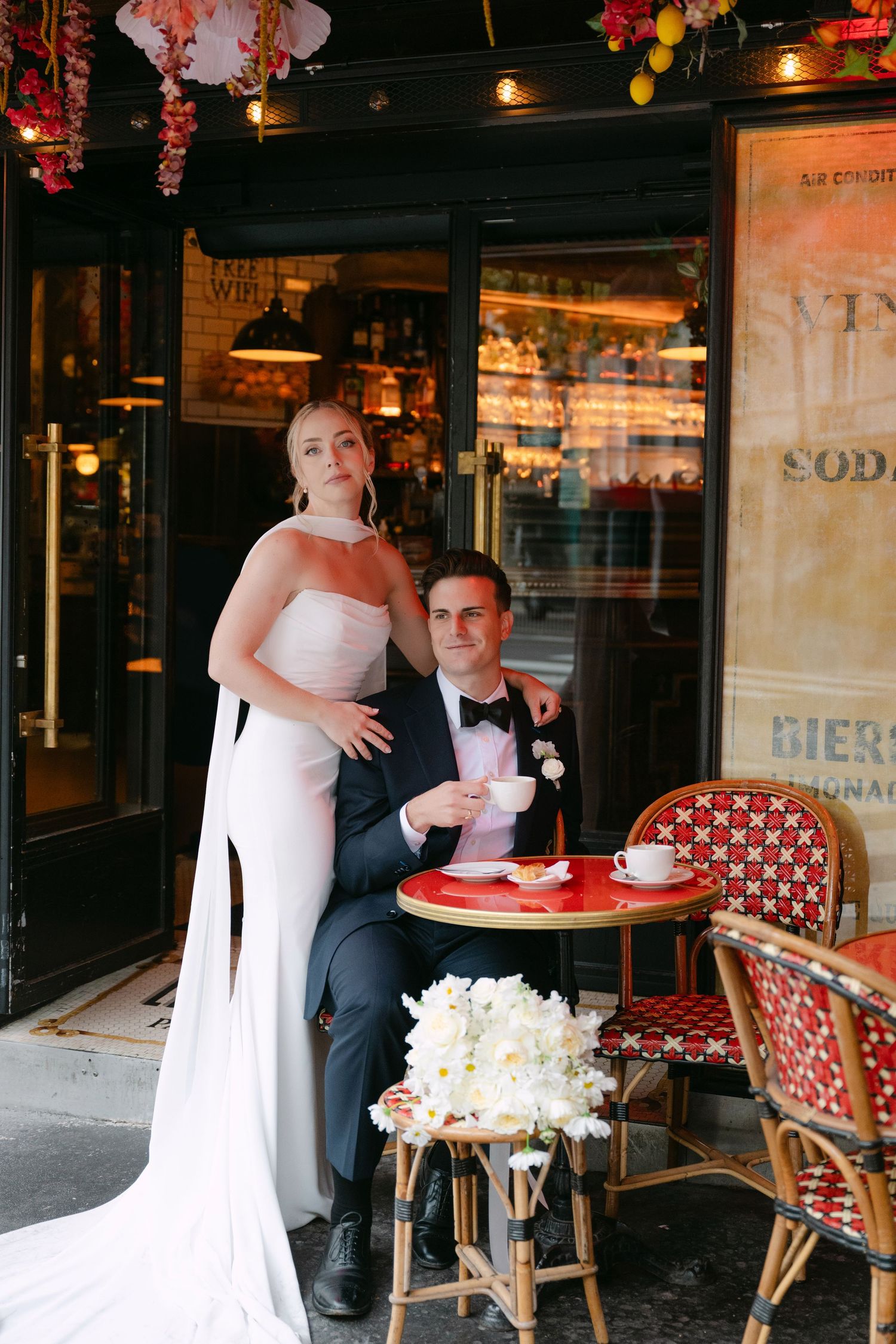 Wedding couple sits at a charming Parisian cafe with red chairs and floral decorations.