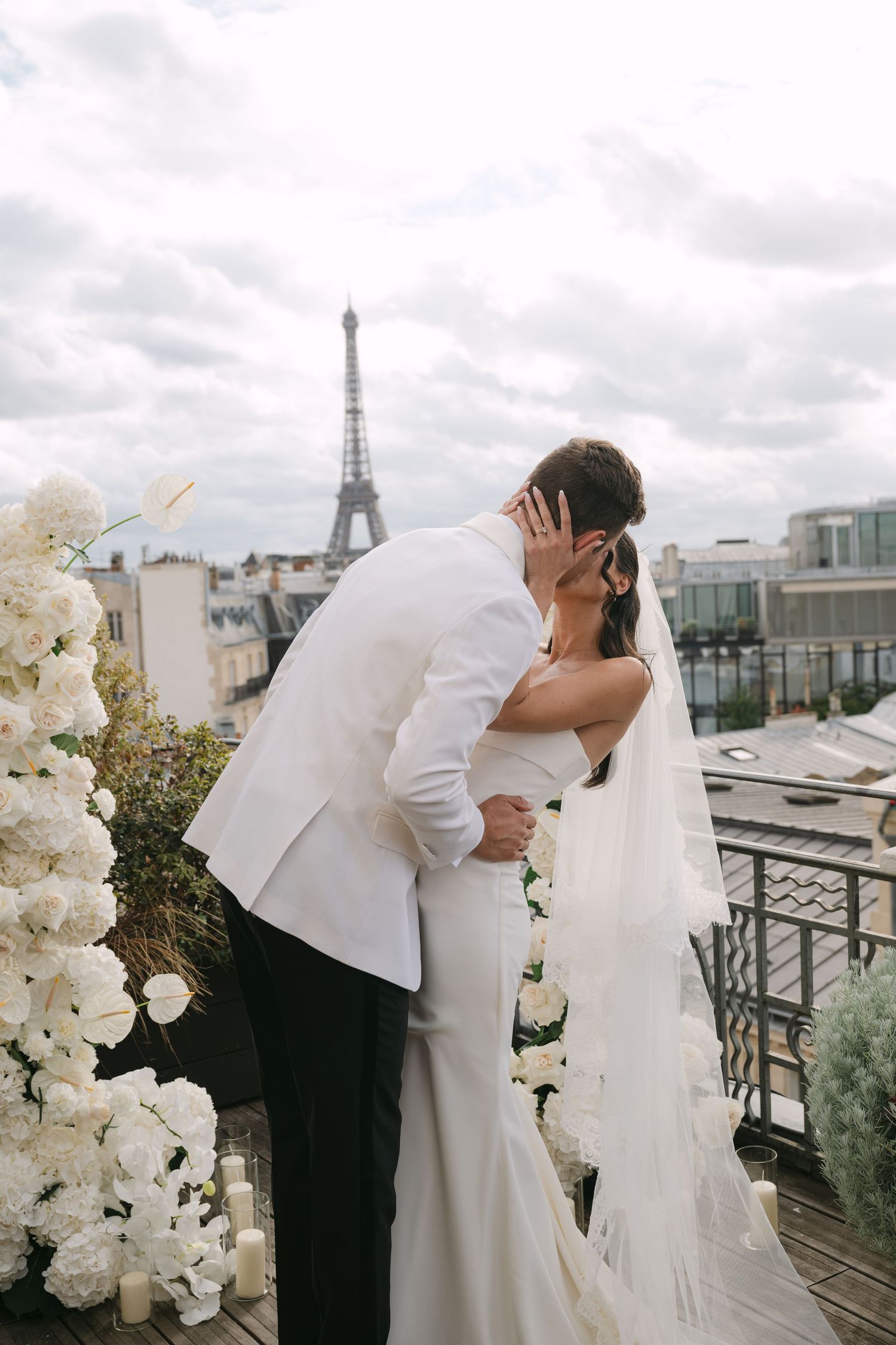 A romantic wedding couple embraces on a Parisian rooftop terrace with the Eiffel Tower visible in the background.