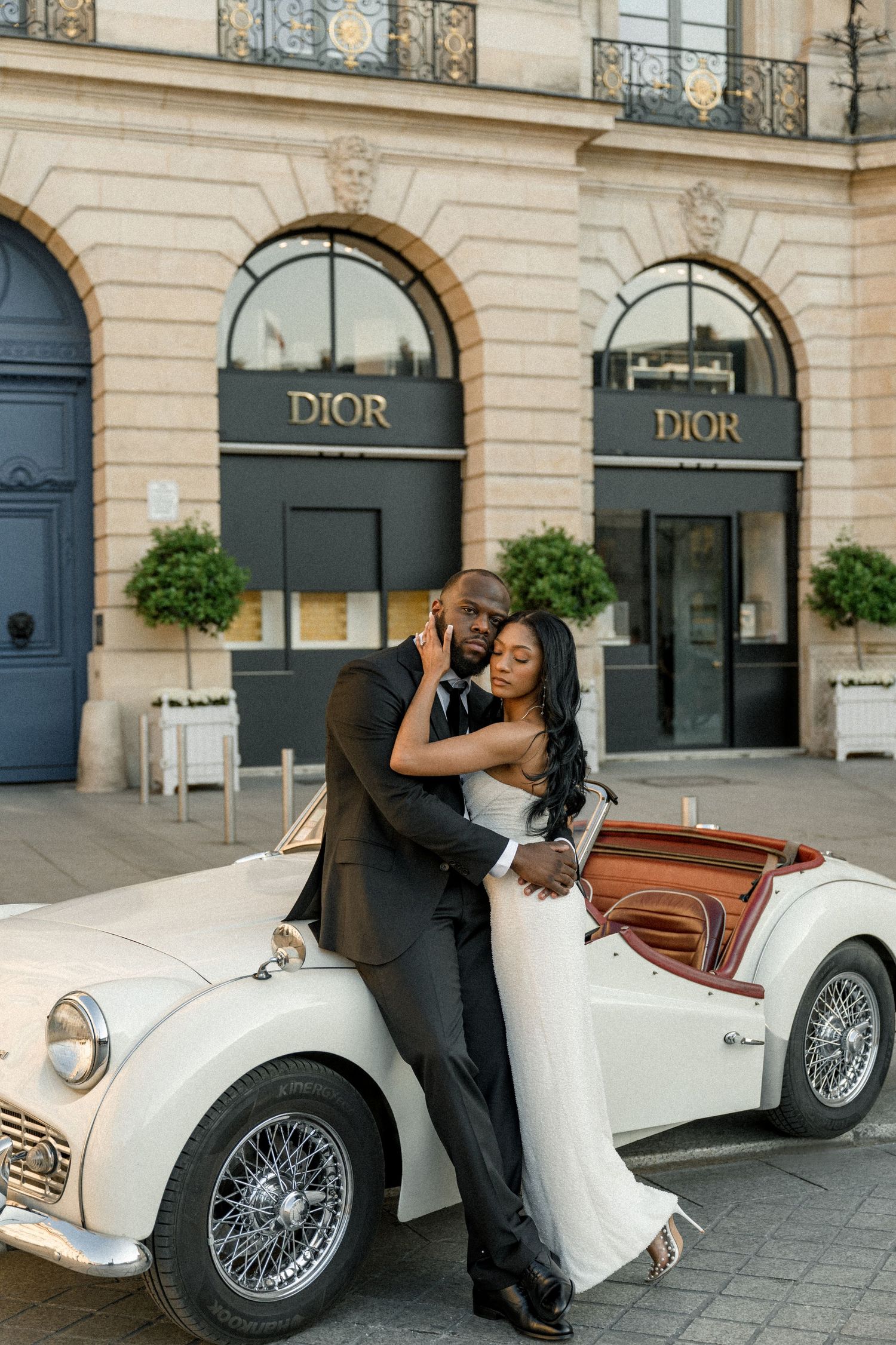 Newlywed couple poses with vintage white sports car outside Dior storefront in Paris.