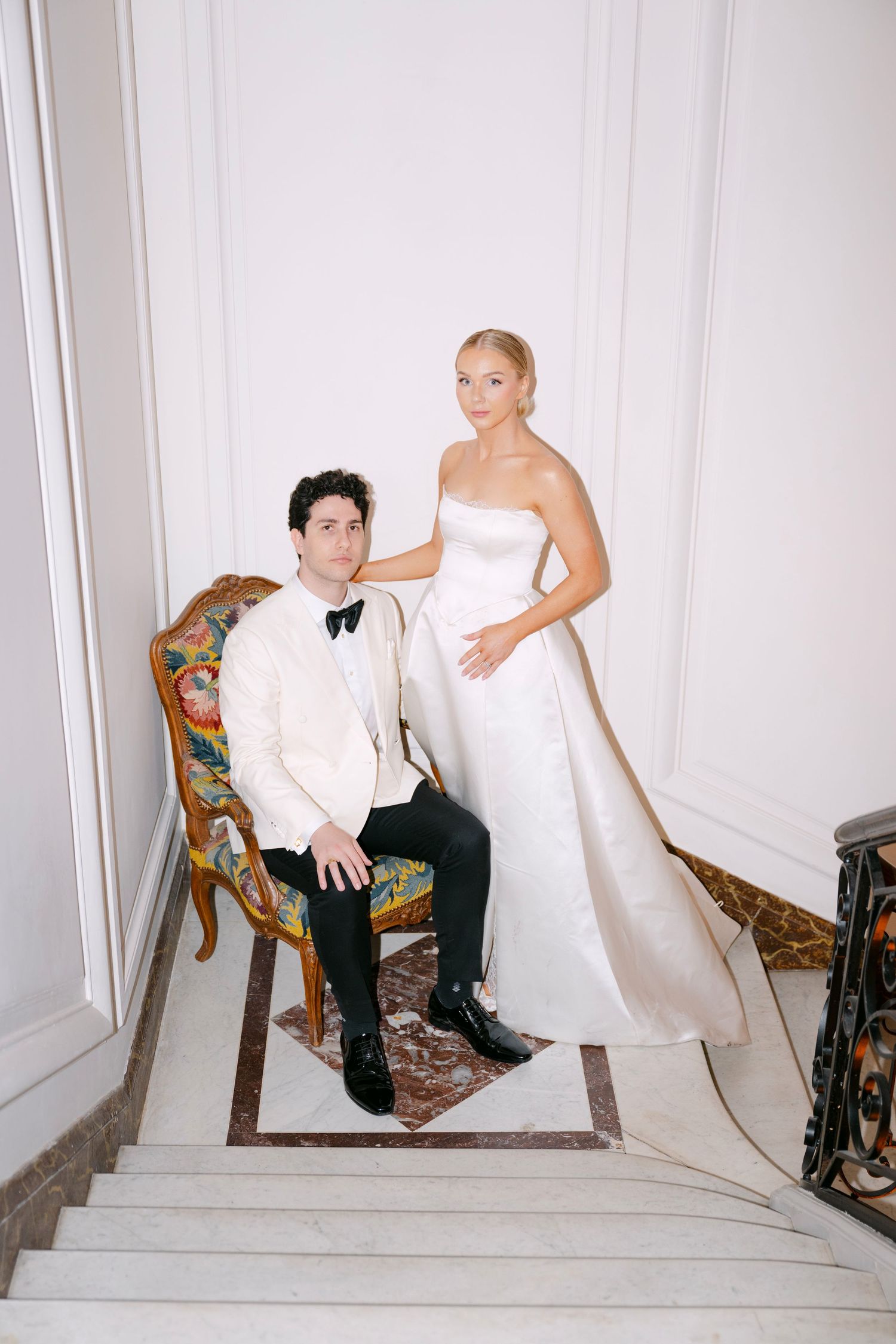 A couple poses on white stairs in a minimalist setting with antique chairs and architectural details.
