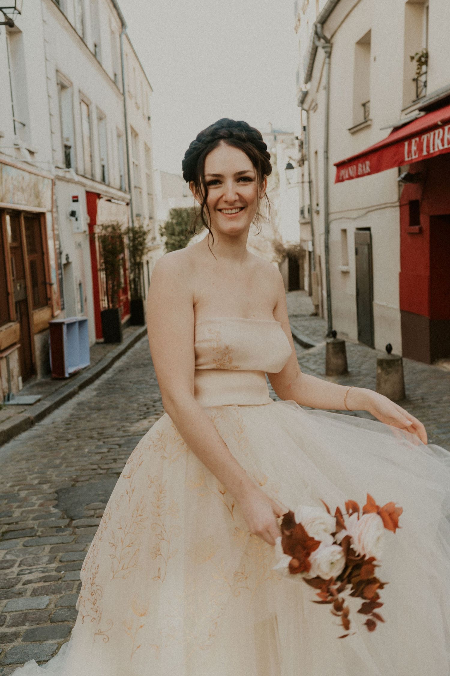 Bride in strapless tulle gown poses on Parisian street holding autumn-colored bouquet.