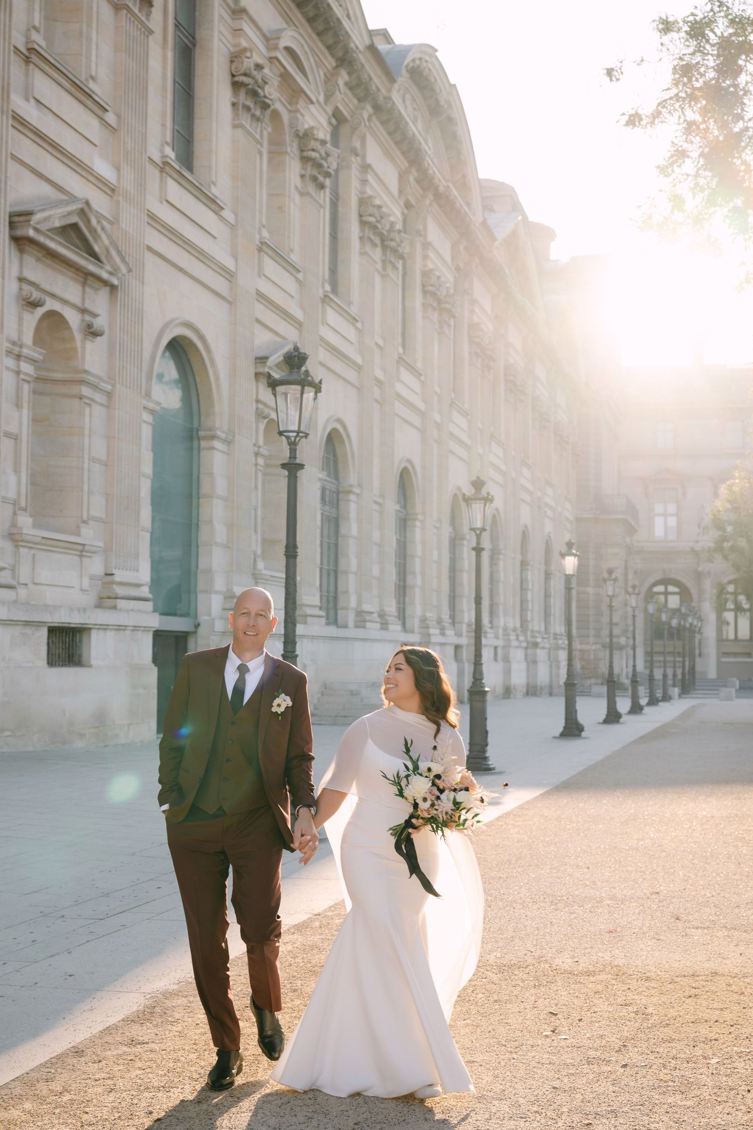 Bride and groom holding hands walking near the Louvre in golden Paris sunlight.