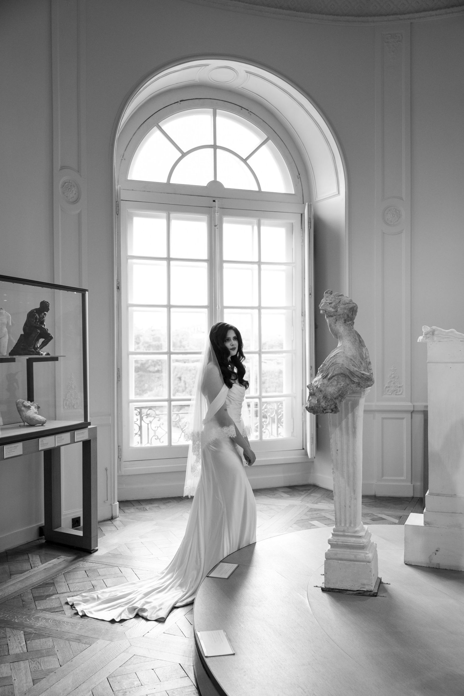 Black and white portrait of bride in flowing satin gown standing by arched window beside classical bust sculpture.