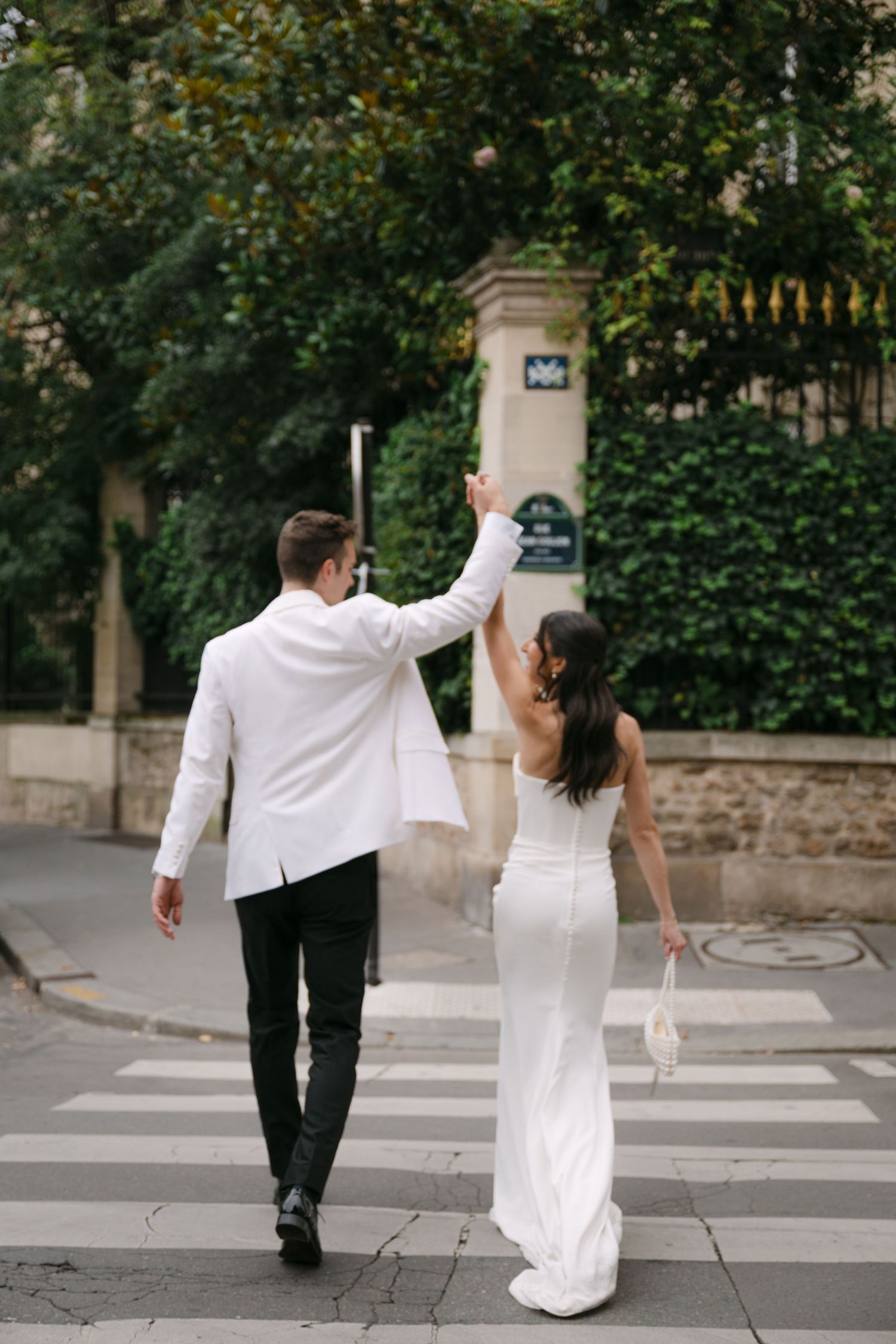 Bride and groom walk across a Parisian crosswalk, raising their joined hands joyfully together.