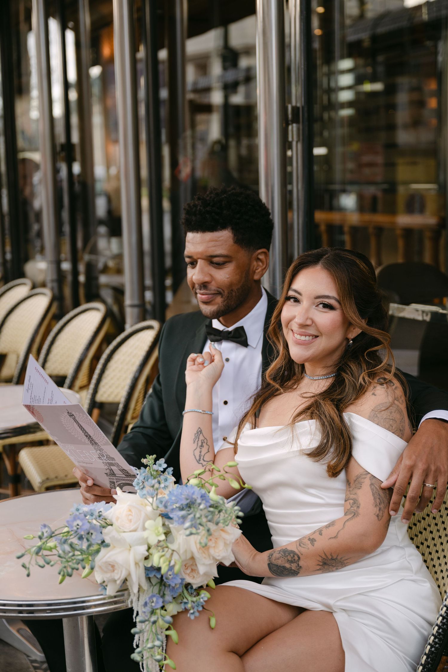 Smiling bride with tattoos and groom in tuxedo reading menus at a classic Parisian café terrace.
