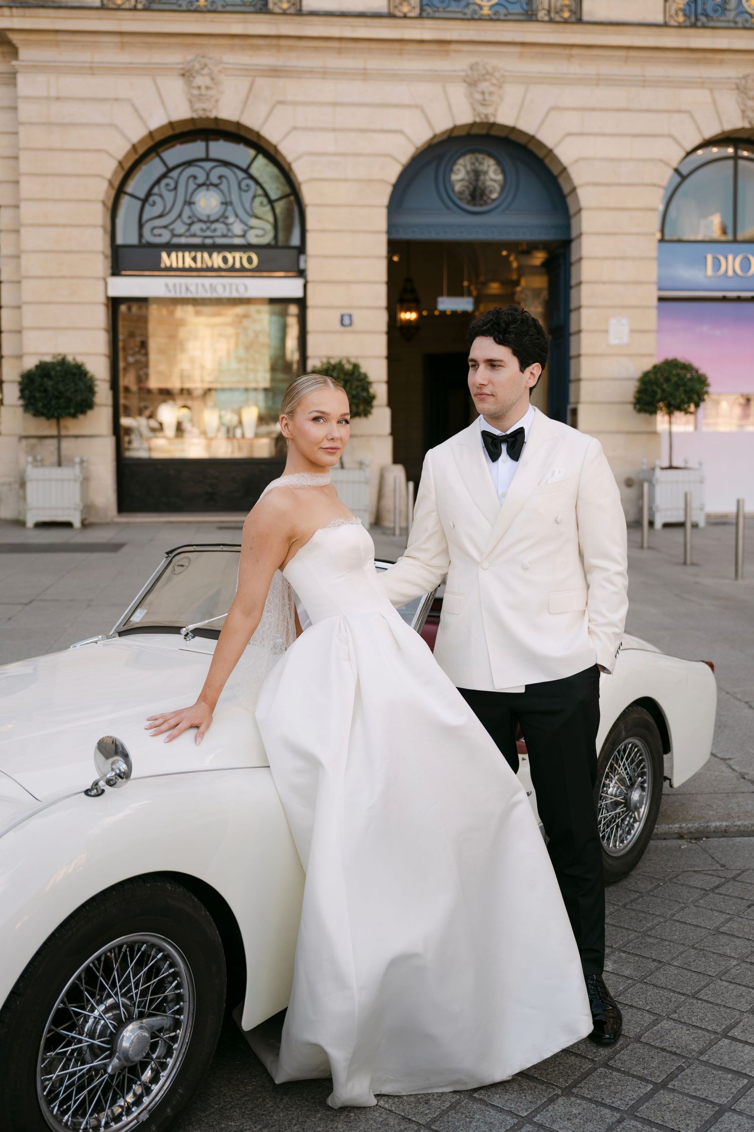 Elegant bride and groom posing beside a vintage white convertible car outside Dior and Mikimoto stores in Paris.
