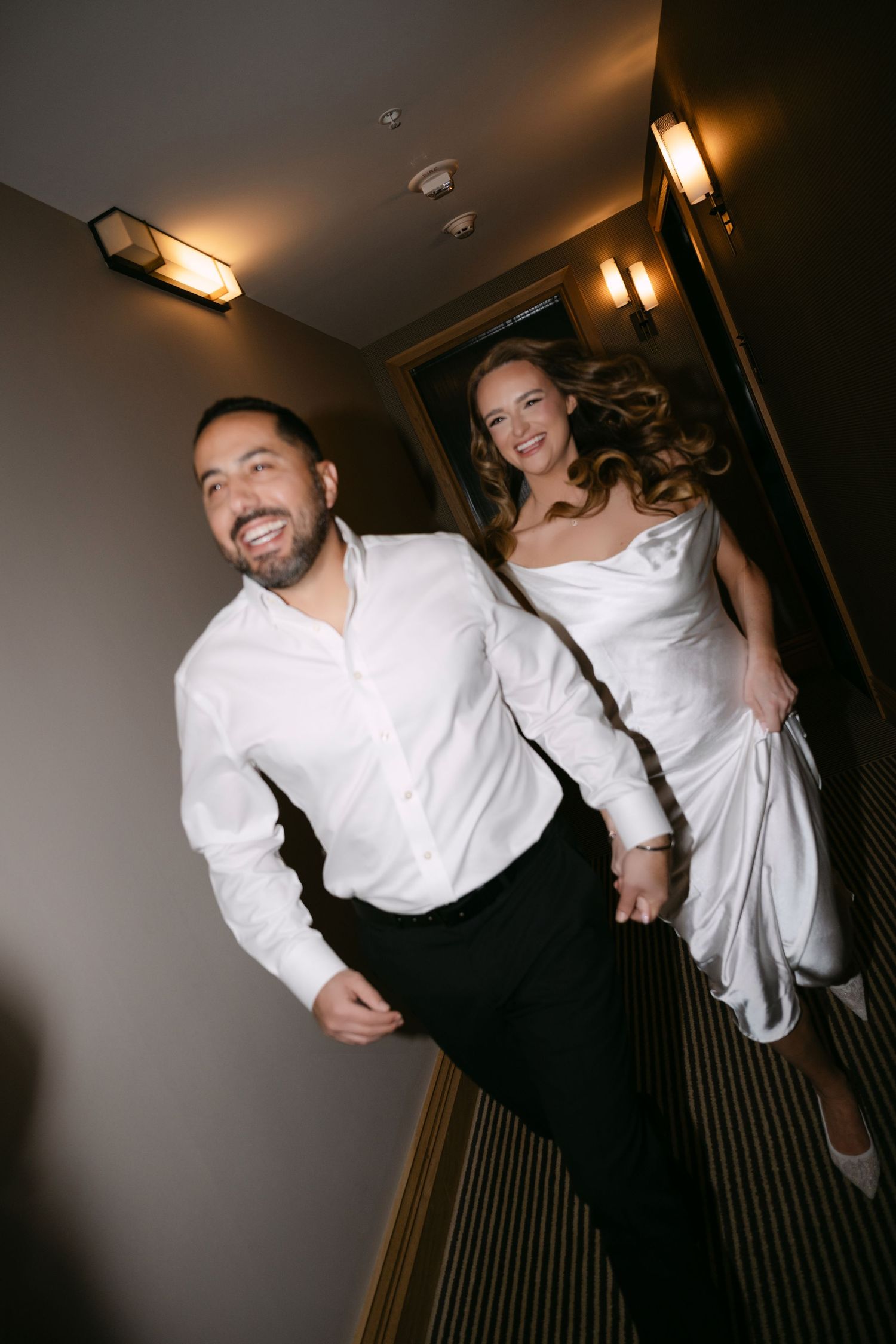 Joyful newlyweds hold hands running through hotel corridor, bride in white gown and groom in white shirt smiling.