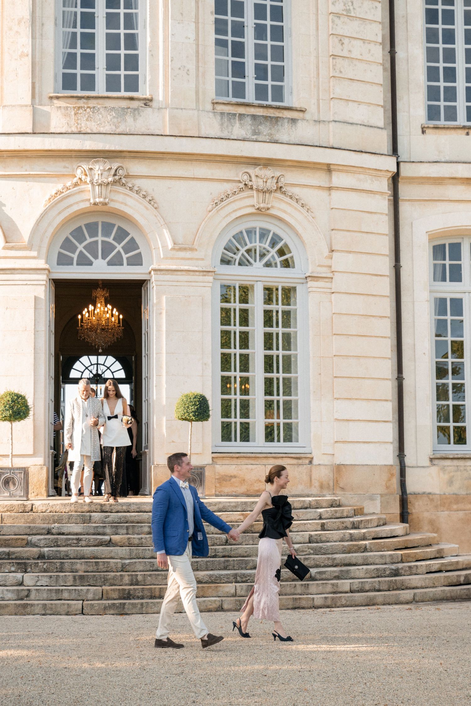 Elegantly dressed couple holding hands walking down steps of a grand French chateau in golden sunlight.
