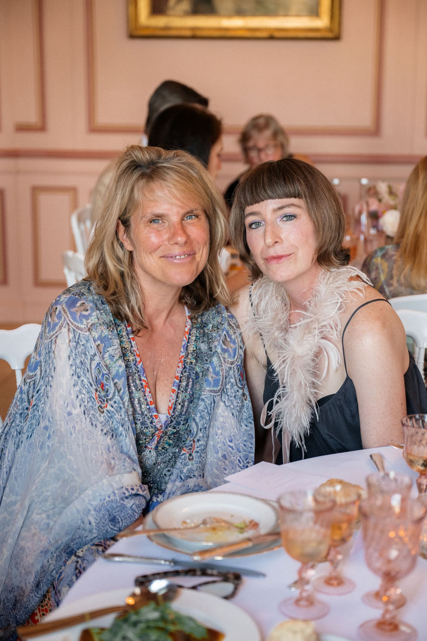 Two women smiling together at an elegant indoor wedding reception dinner in a pink-walled venue.