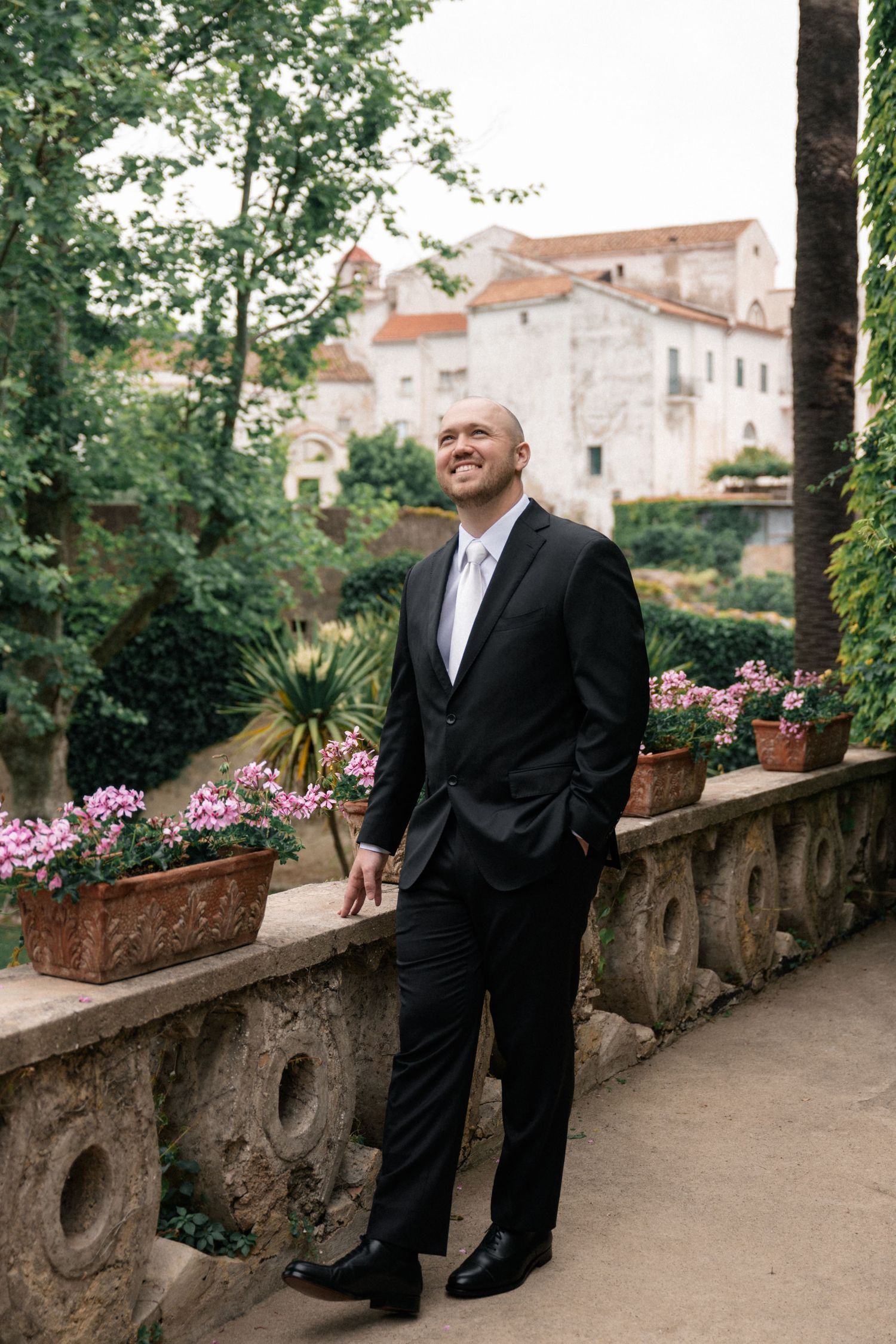 Smiling groom in black suit standing in Italian garden with pink flowers and historic building.