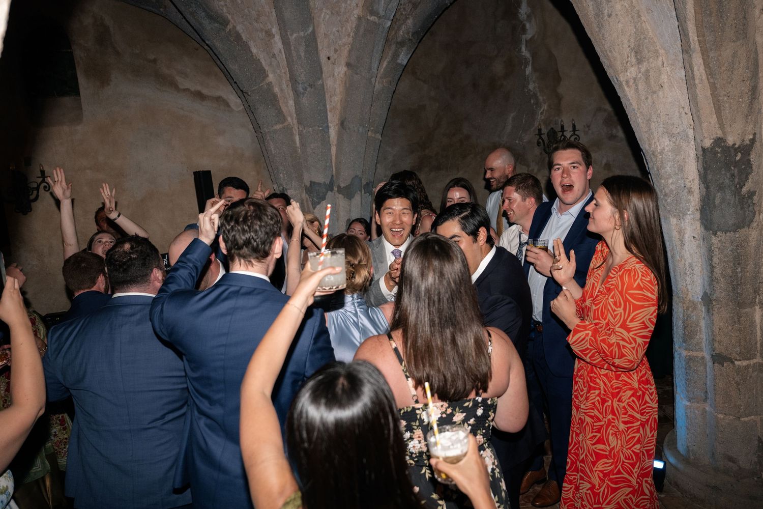 Wedding guests celebrate and dance joyfully under stone arched ceilings at a rustic Italian venue reception.